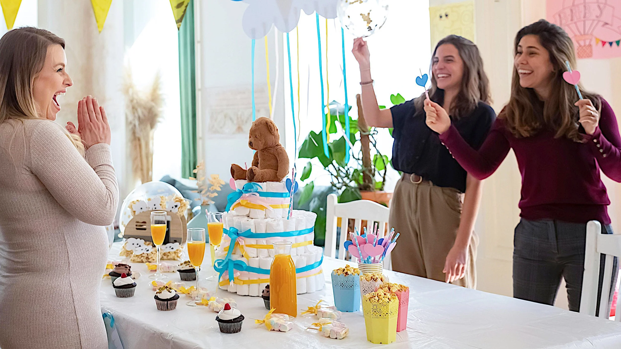Happy Pregnant Mom Celebrating a Baby Shower With Guests. Table is Decorated With Party Favors, Treats, and a Diaper Cake Topped With a Toy Teddy Bear.