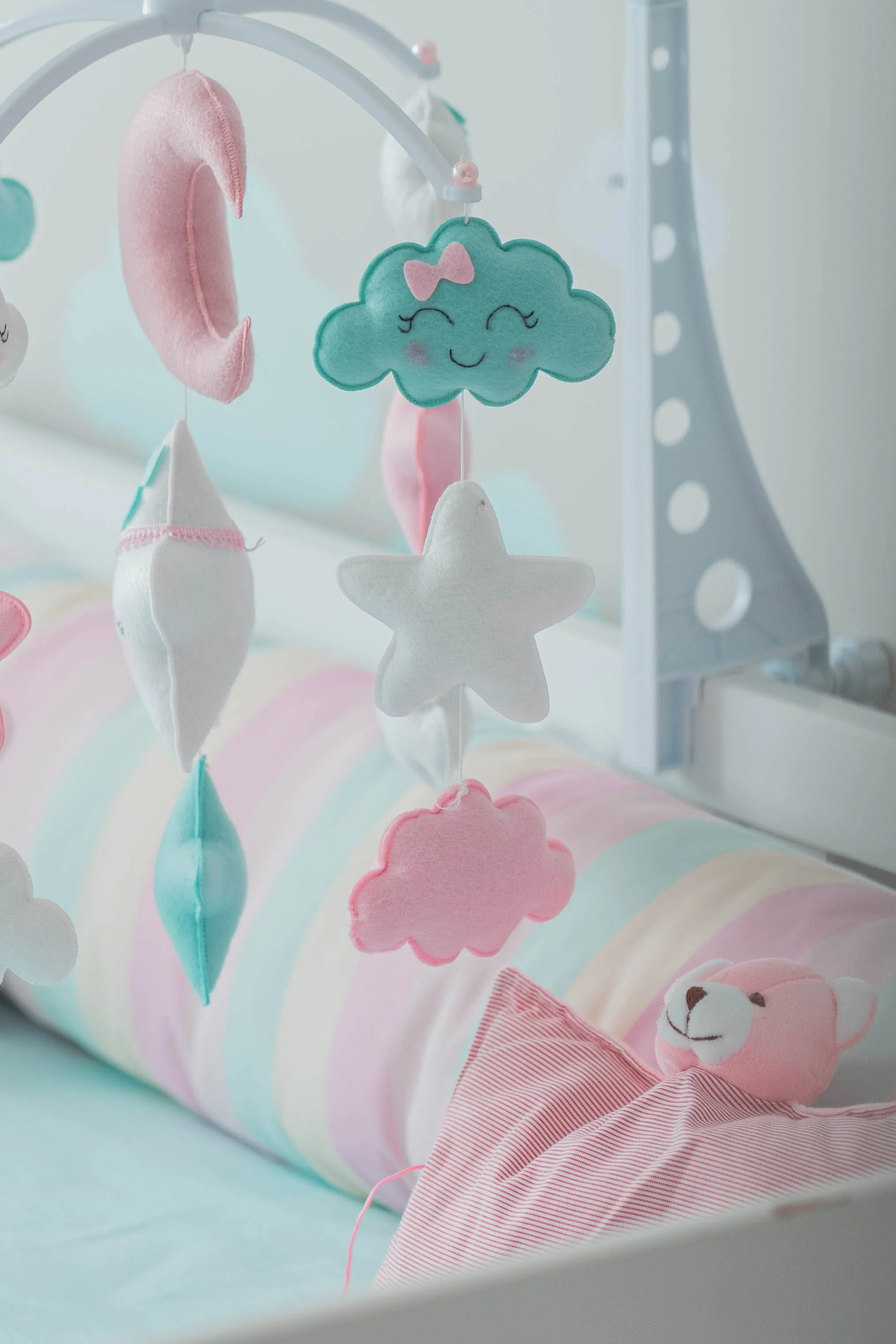 Baby Nursery Crib Decorated With Bumper, Mobile, and Toy Teddy Bear in White and Pastel Colors of Pink, Blue, and Yellow.