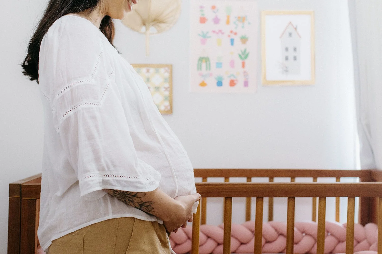 Smiling Pregnant Mom Standing in Baby Nursery With Hands Cradling Her Baby Bump. Pictures Decorate the Wall and Crib Has A Pink Bumper.