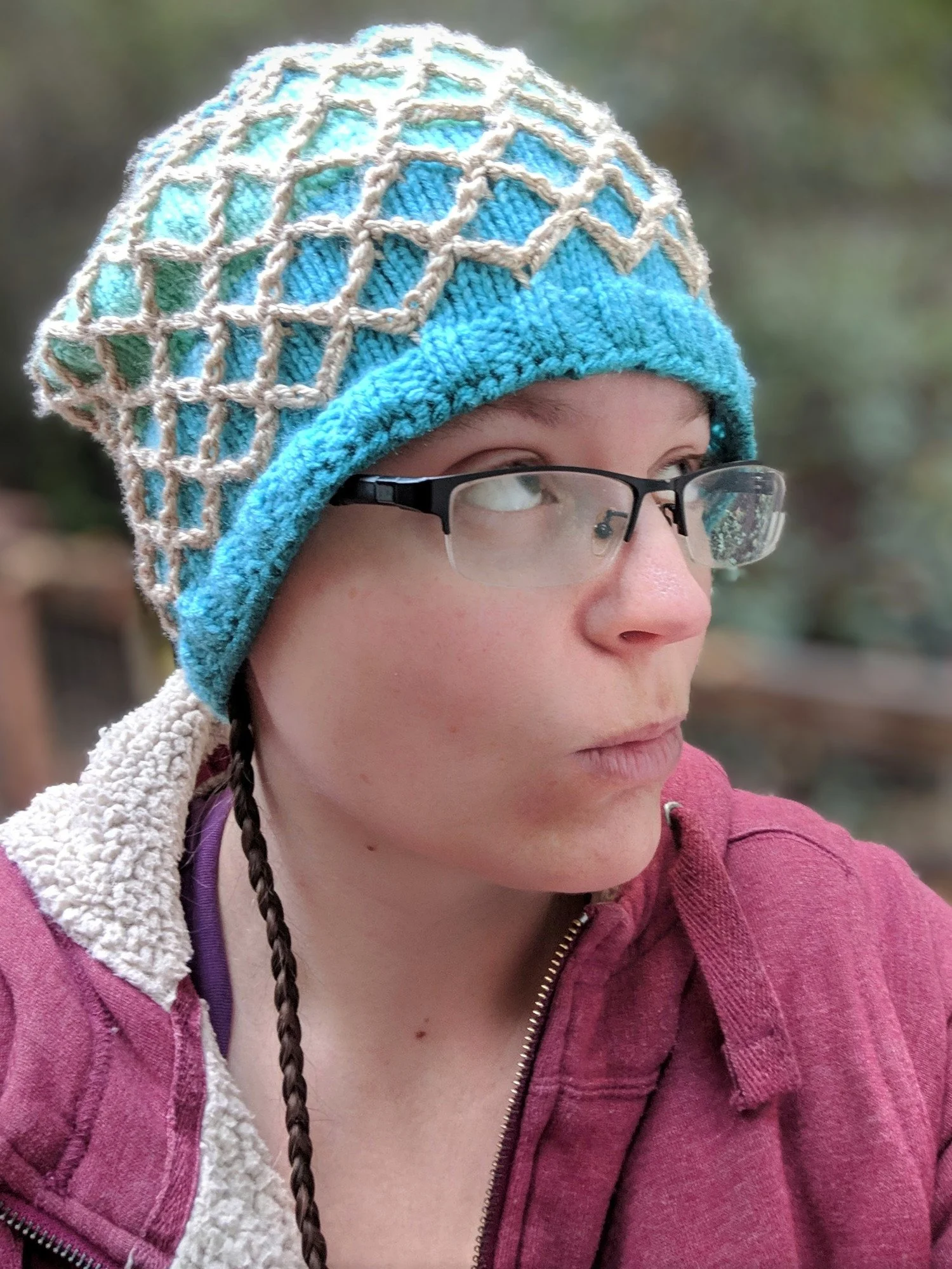 A person with glasses wearing a colorful knit hat and a pink hoodie, outdoor setting