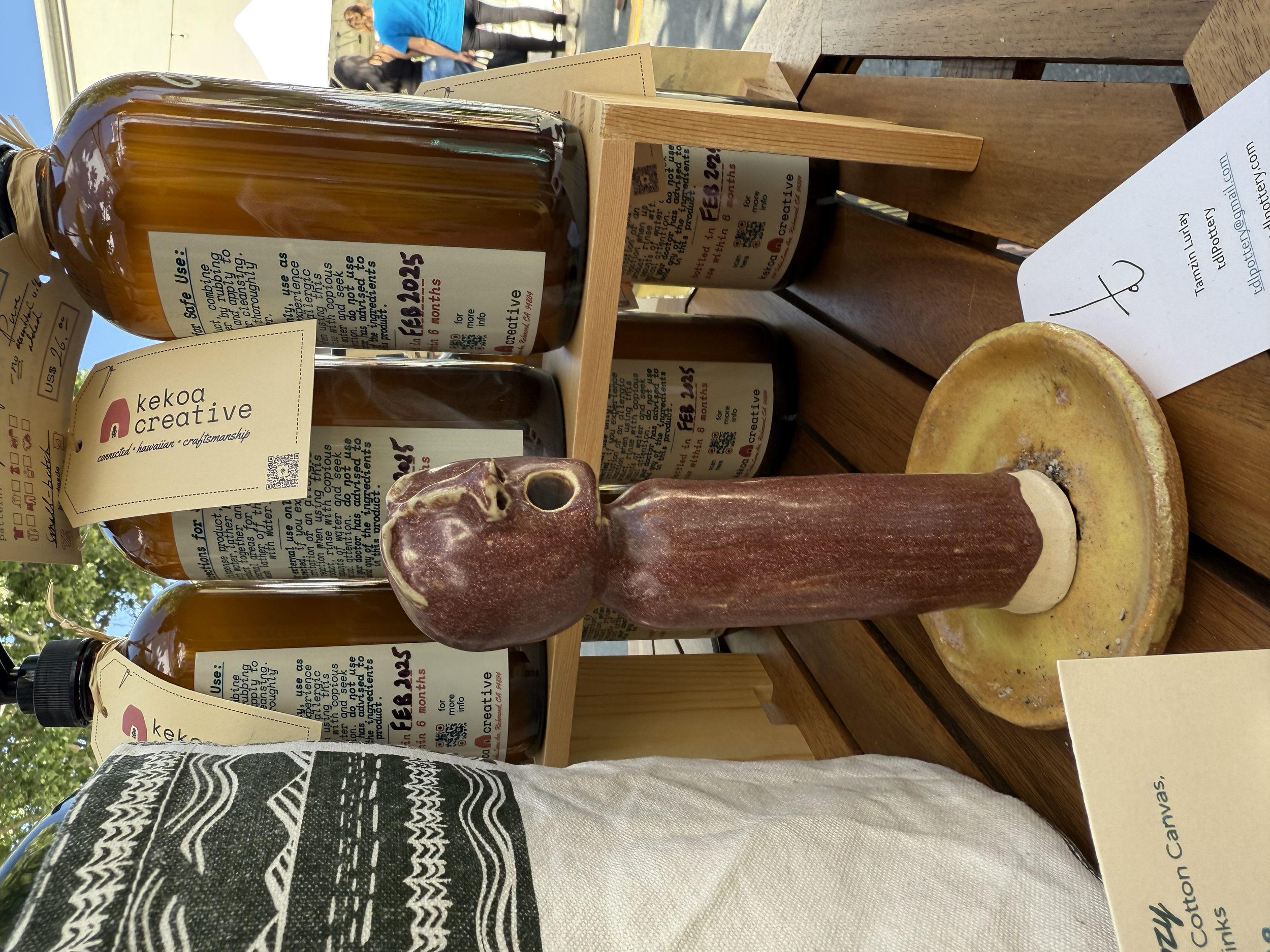 Display of Kekoa Creative products, including bottles of thick liquid and a clay or ceramic sculpture on a wooden table, with some printed labels and price tags.