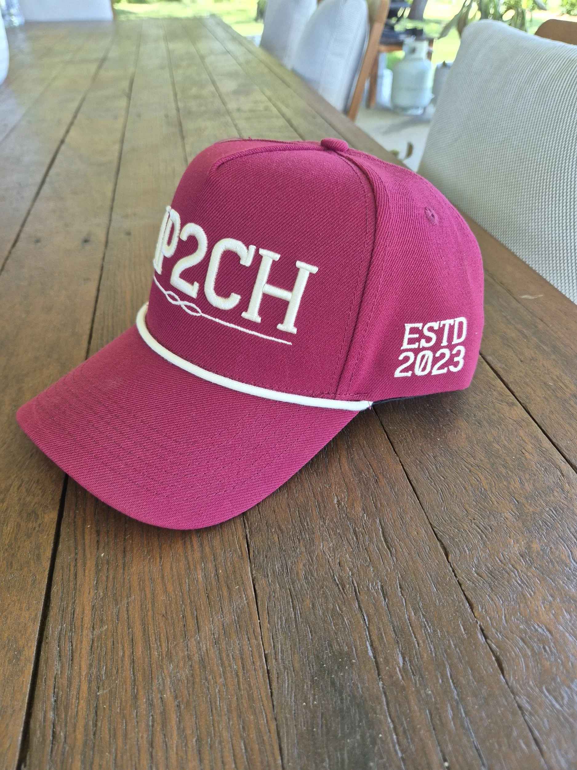 Pink baseball cap with white embroidered text reading '20' and 'CH' and 'ESTD 2023' on the side, placed on a wooden table.