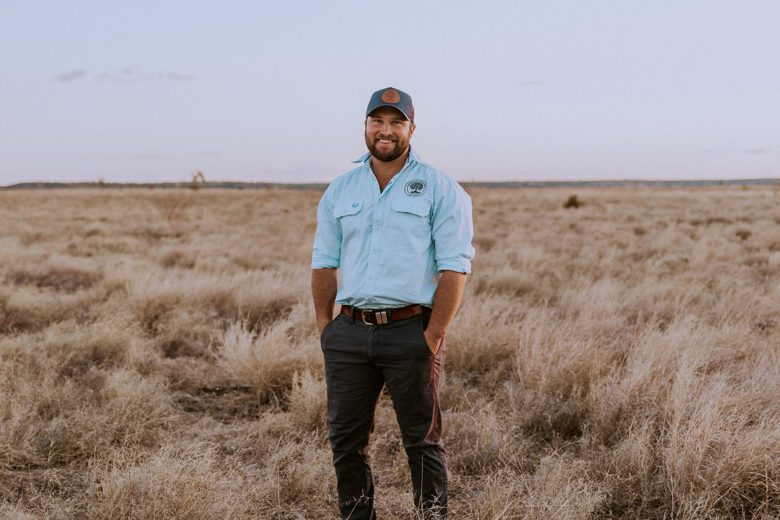 Man in light blue outdoor shirt and dark pants standing in dry grassland with a horizon and cloudy sky in the background.