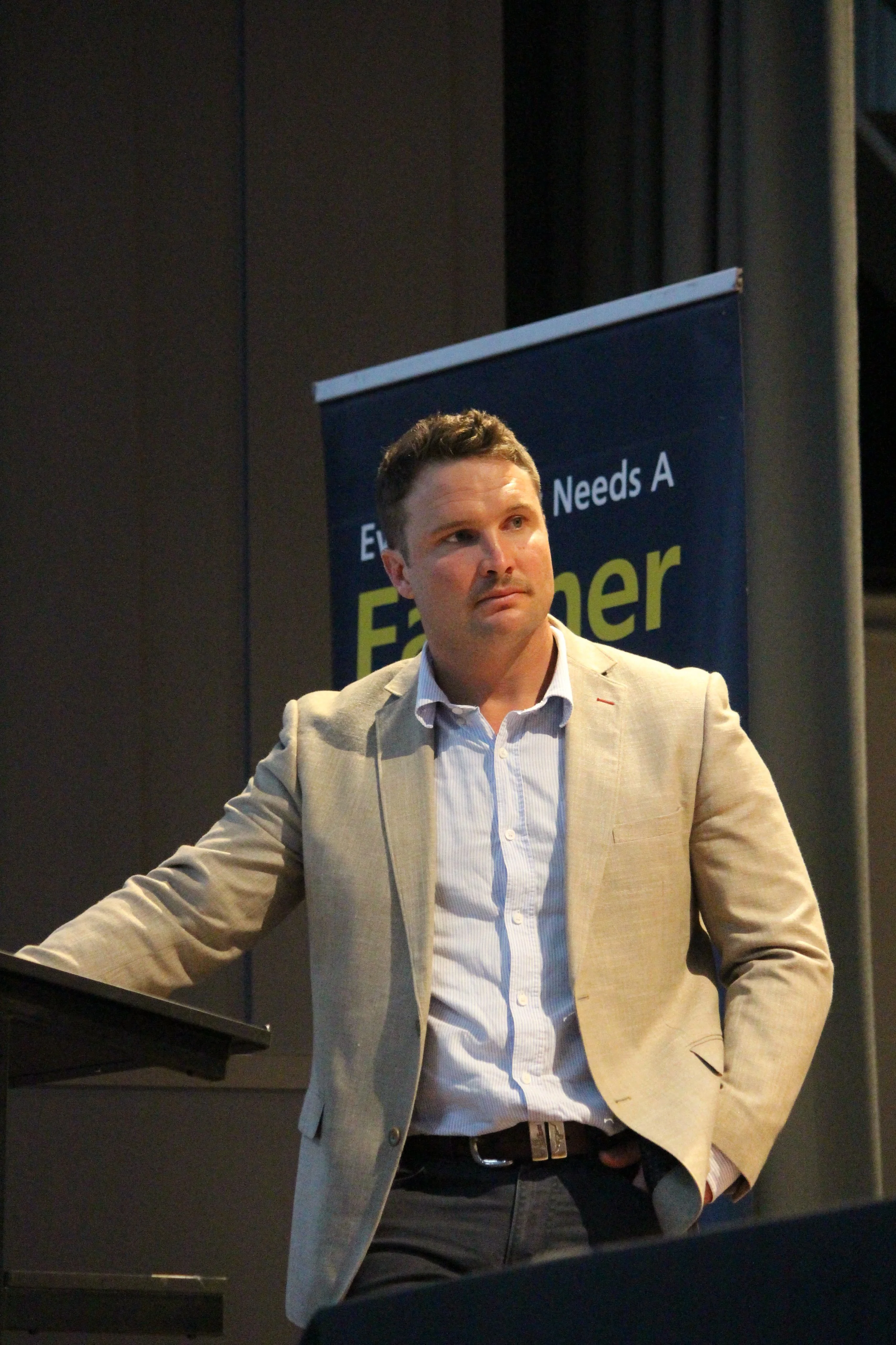 A man in a beige blazer standing in front of a dark background with a blue sign that says "Everyone Needs A Farmer".