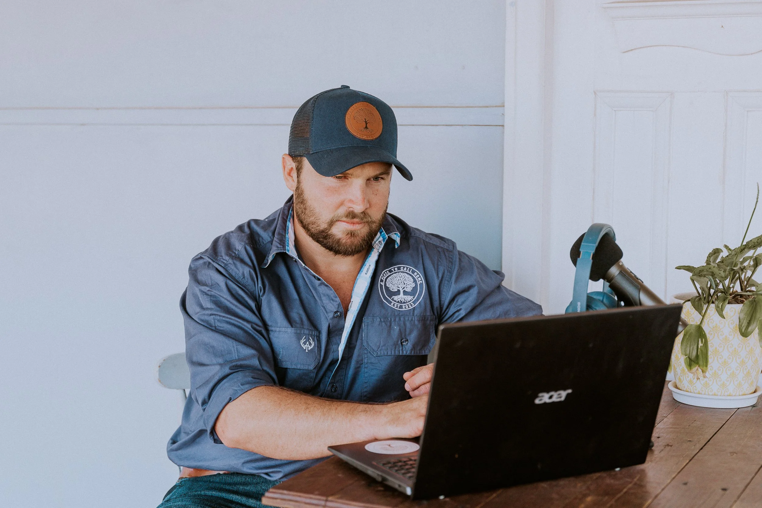 Man sitting at a wooden table, wearing a blue cap and shirt, using a black Acer laptop, with a pair of headphones and a potted plant nearby.