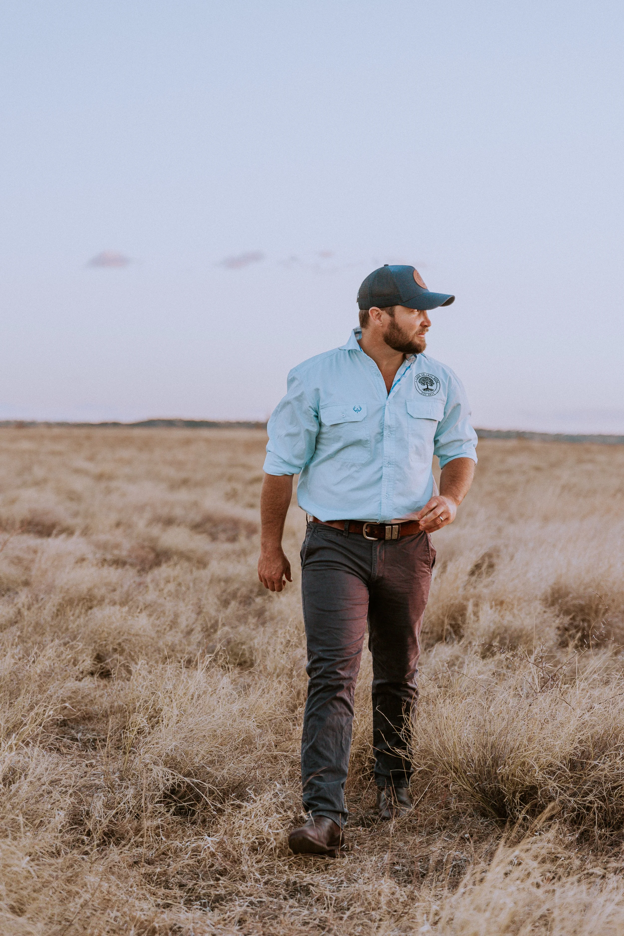 A man walking through a grassy field at dusk, wearing a light blue long-sleeve shirt, dark pants, a belt, boots, and a black cap, with no visible animals or objects.