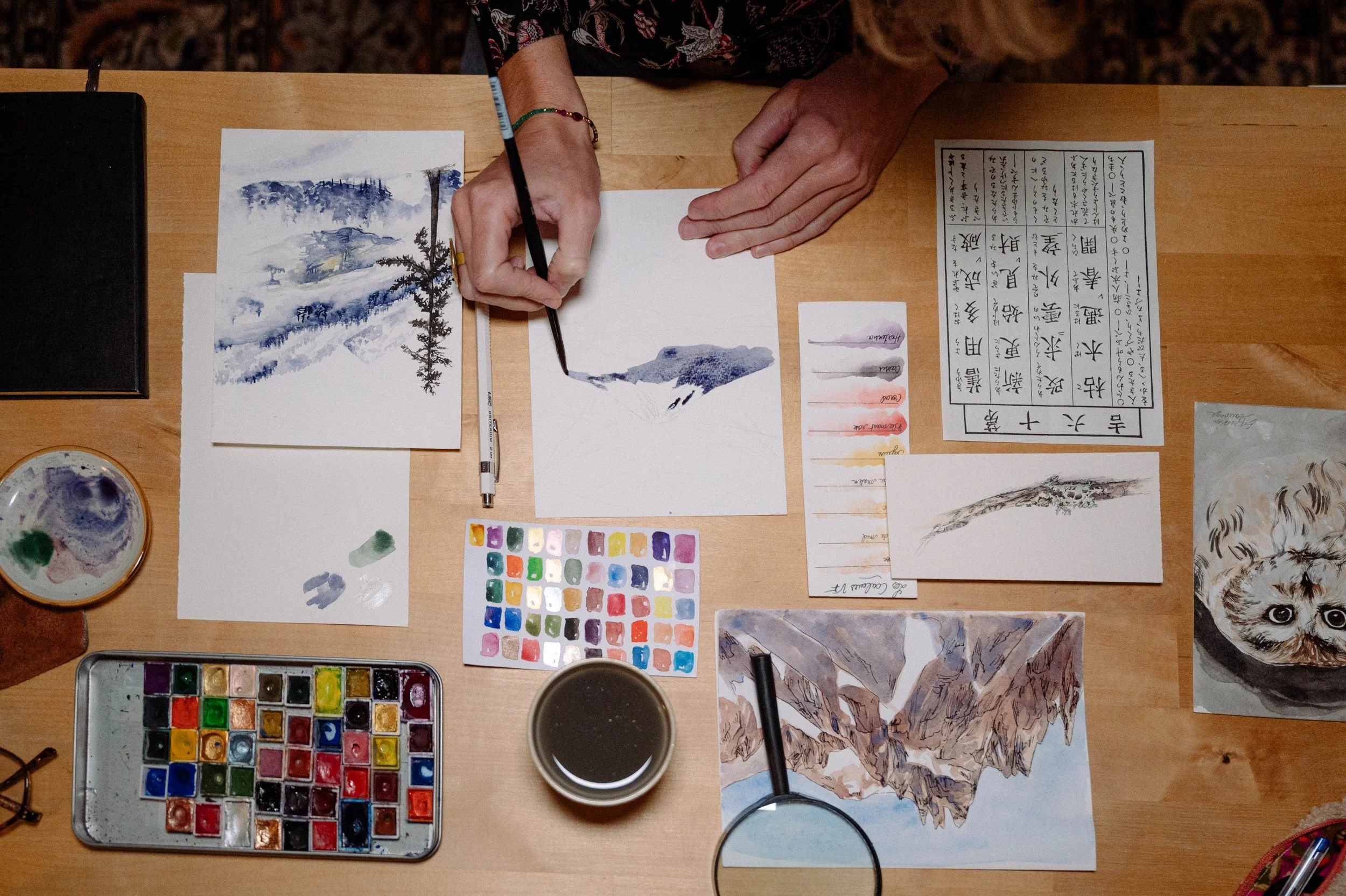 Expression Sauvage peignant une scène de montagne avec de l'aquarelle, entourée de diverses couleurs, pinceaux, et matériaux de peinture sur une table en bois.