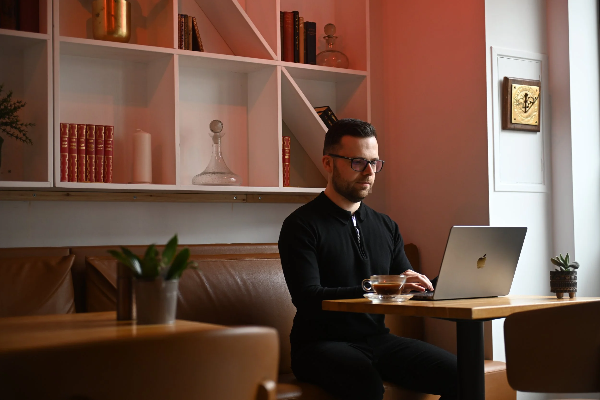 A man with glasses and a beard working on a silver laptop at a wooden table in a cozy cafe. The table has a cup of coffee, and there are a small potted plant and a larger plant on the window sill. The background features a pink wall, white shelves with books and glass decanters, and a framed clock.