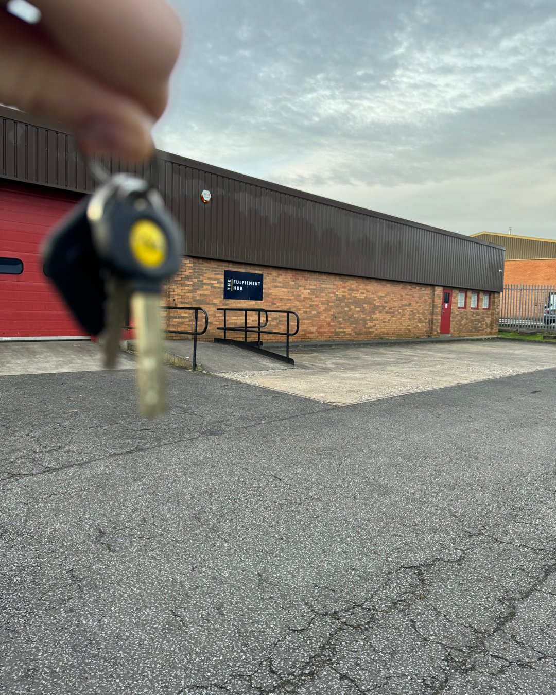 A person's hand holding a car key with a yellow logo, in front of a building labeled 'The Fulfilment Hub.' The building has a brick lower section and a dark upper section, with a red door and a wheelchair ramp outside.