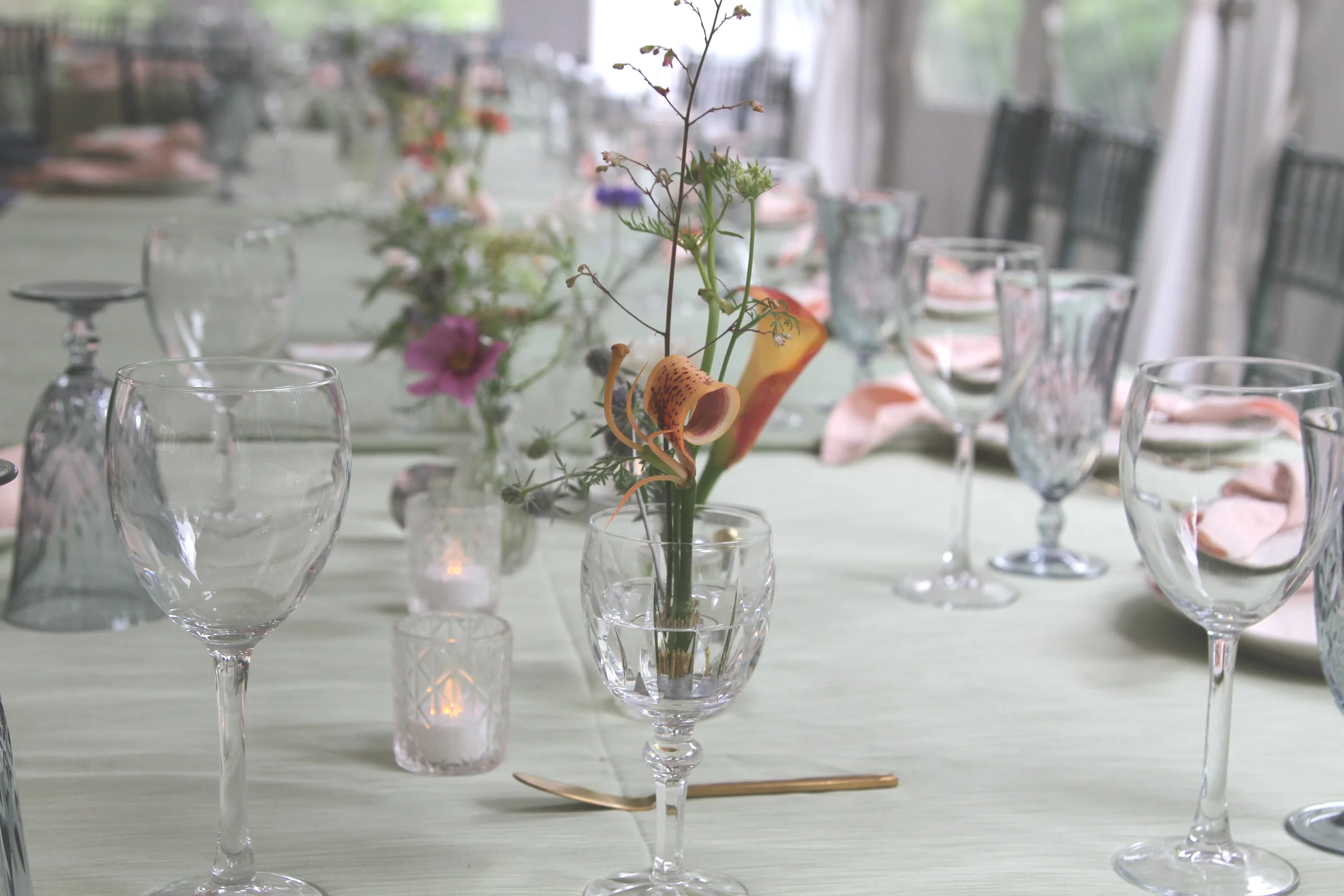Elegant dining table with multiple empty wine glasses, a floral centerpiece, candles, and scattered pink rose petals.