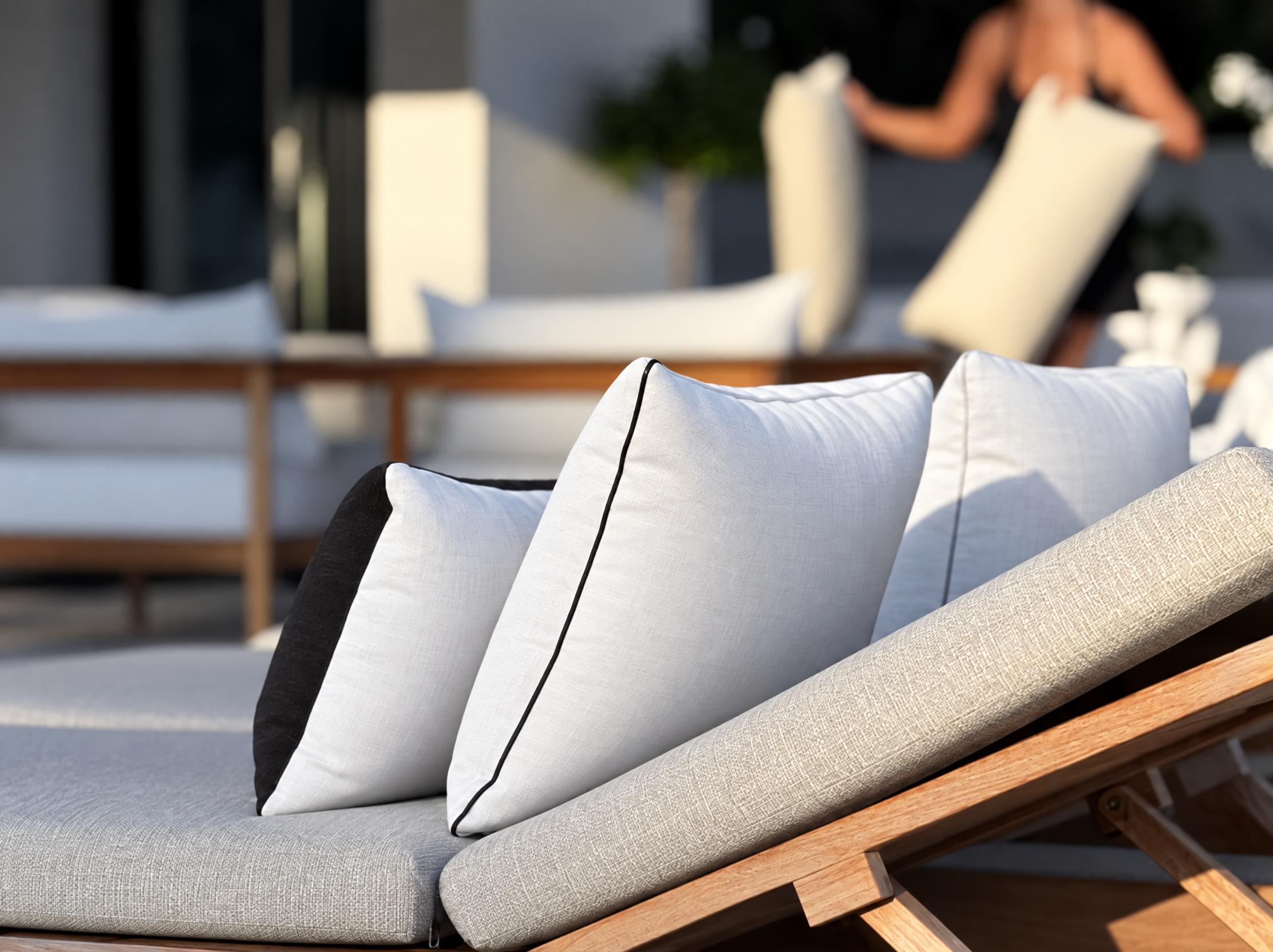 Outdoor seating area with beige cushioned lounge chairs on a wooden frame, decorated with white pillows with black piping, illuminated by sunlight.