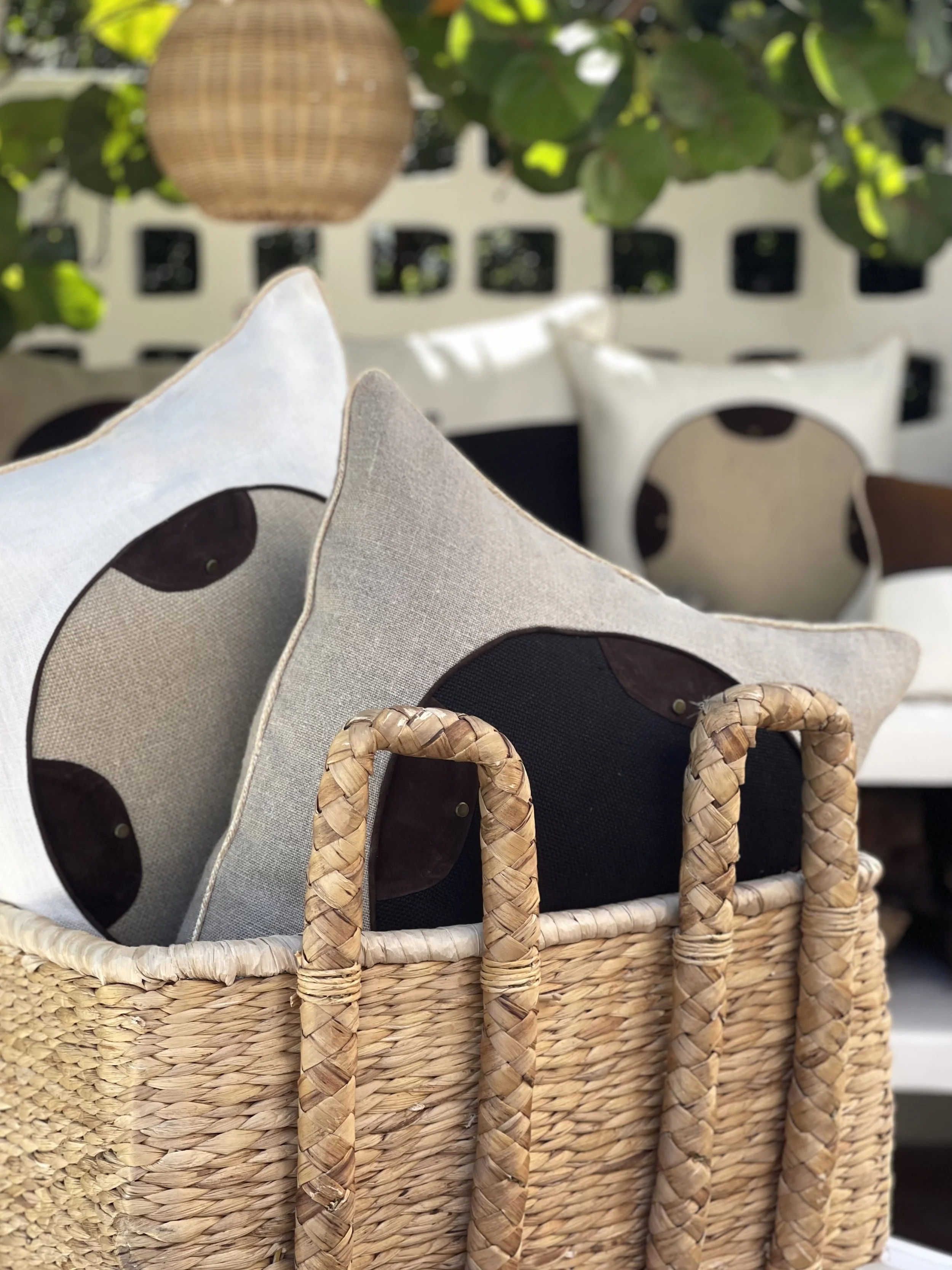 Close-up of a woven basket with Palme Bandero pillows inside, featuring black circle center, with blurred green leaves and a beige wall in the background. Palme Bandero Pillows.