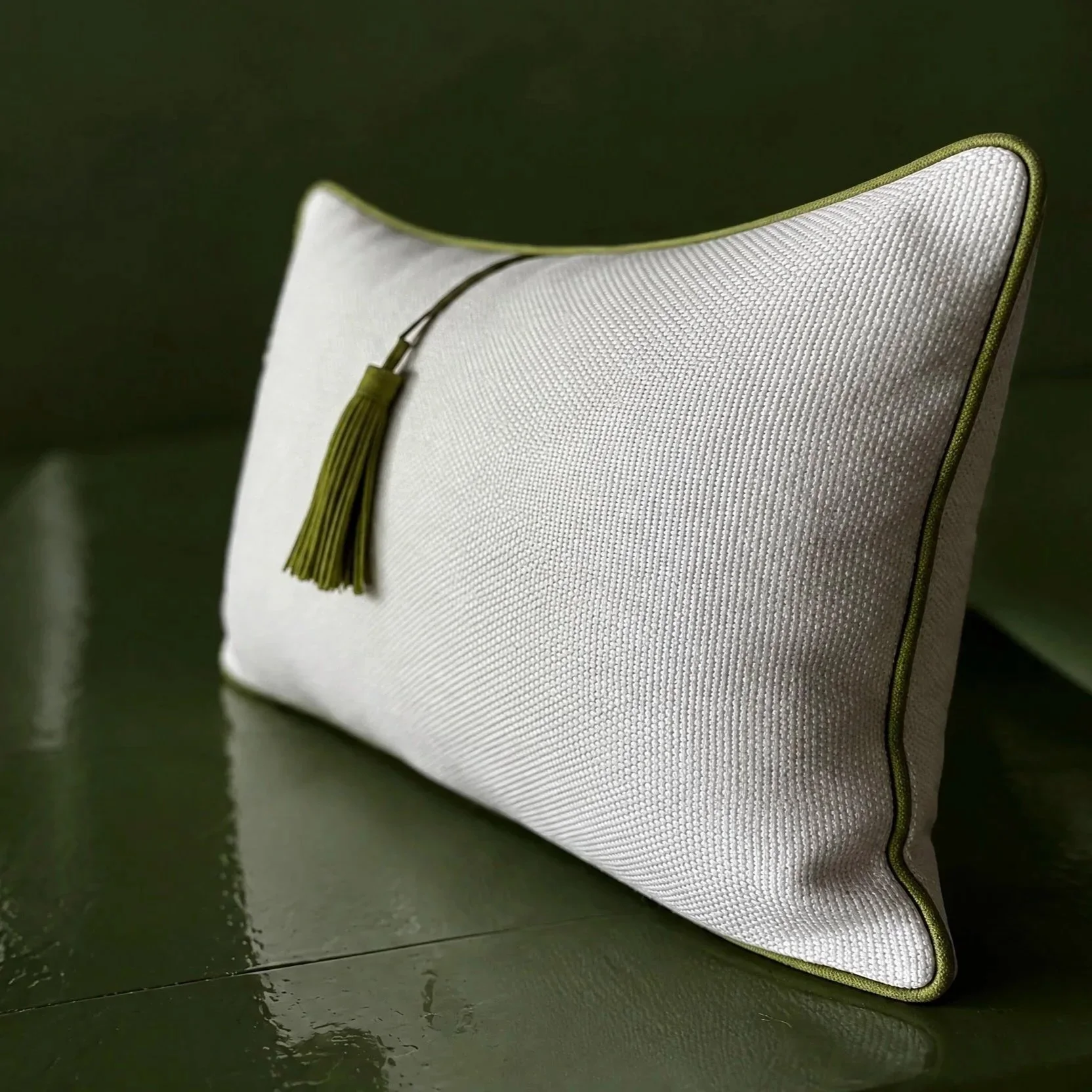 Close-up of a white textured pillow with green piping and a tassel on a dark green surface.