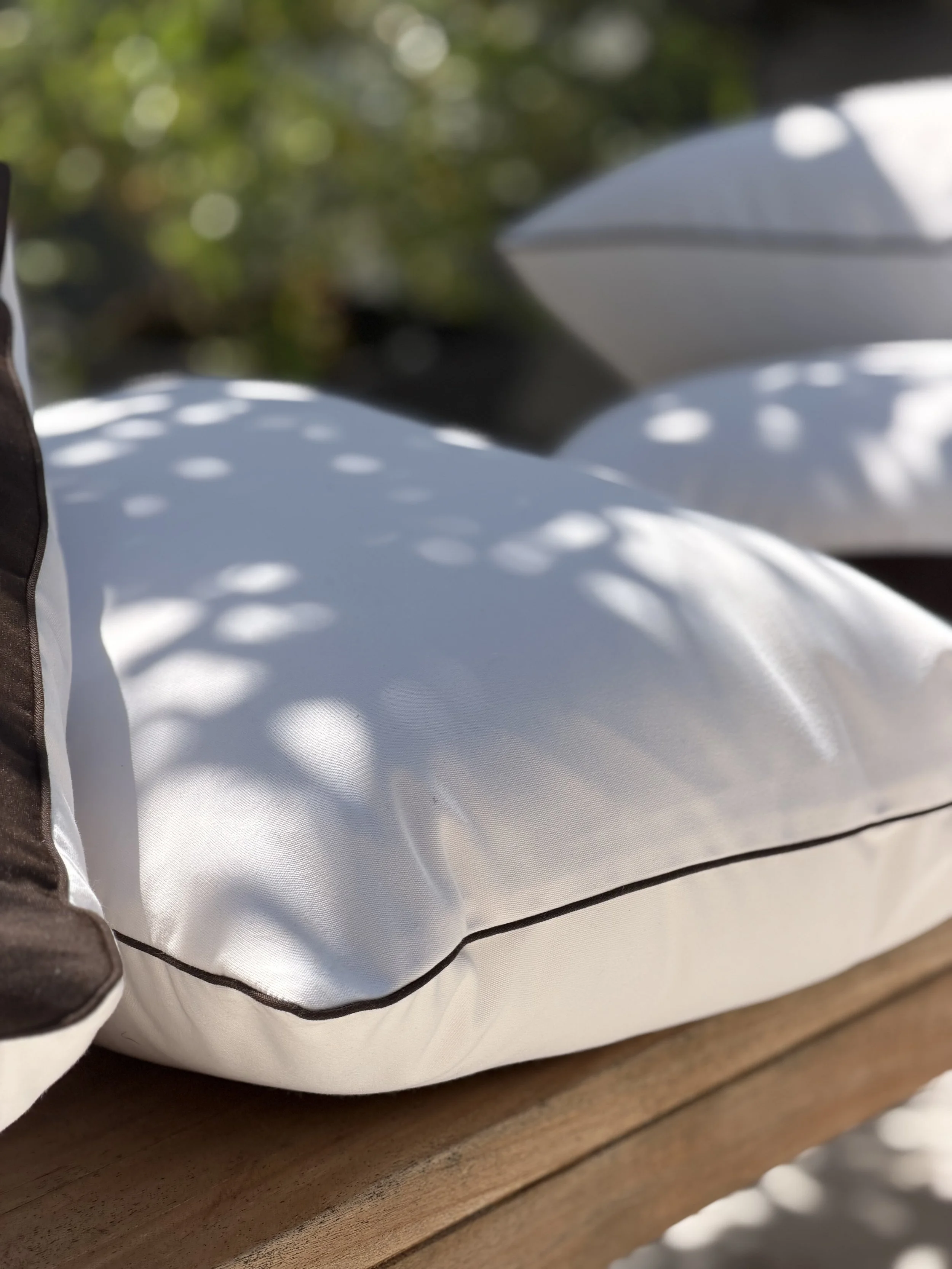 Close-up of a outdoor pillows with white fabric and  a brown outline, shadows created by light and leaves, and a dark edge on the left.