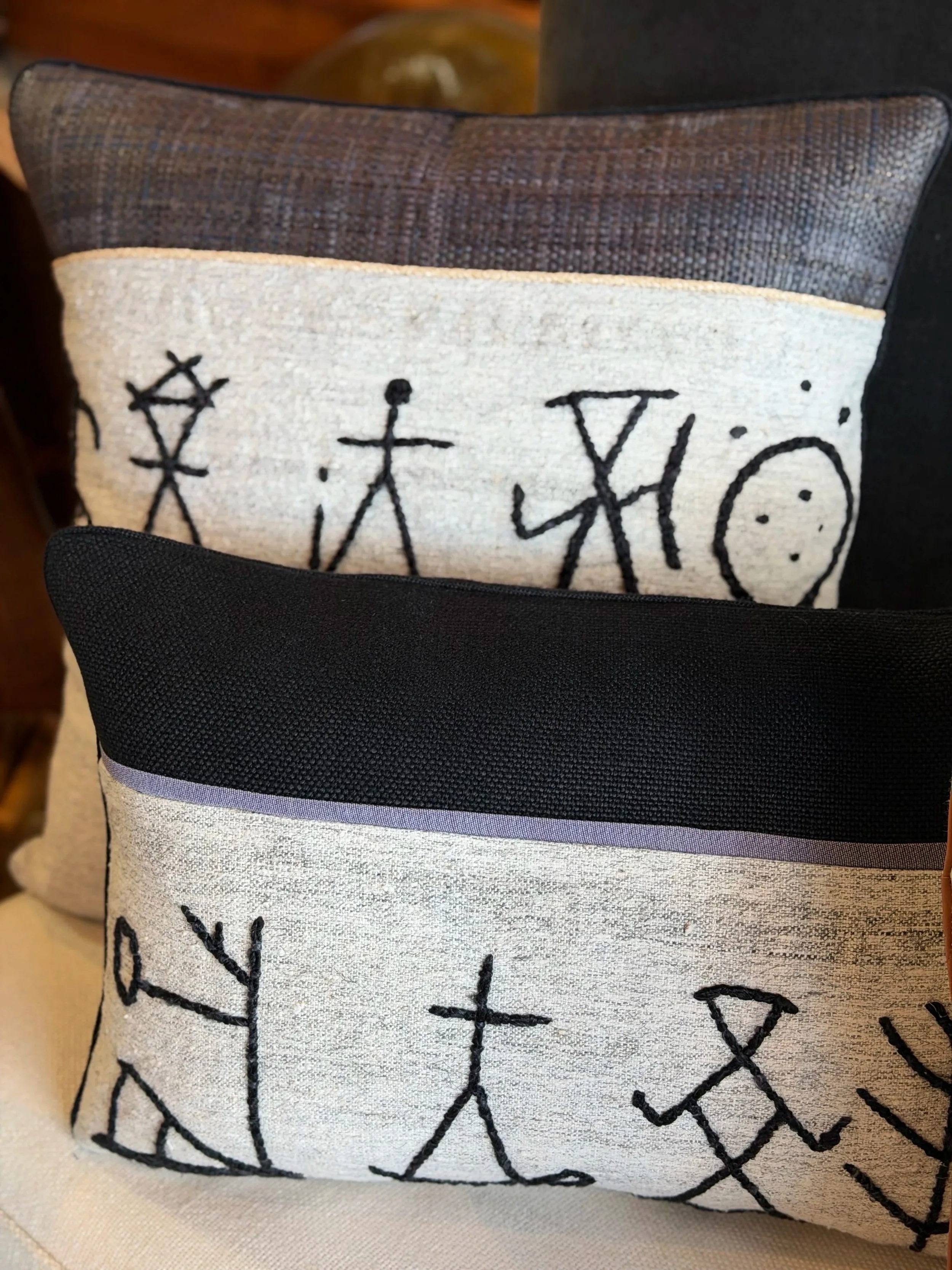 Close-up of decorative cushion with embroidered black symbols on white fabric, with a dark textured cushion and other textiles in the background.