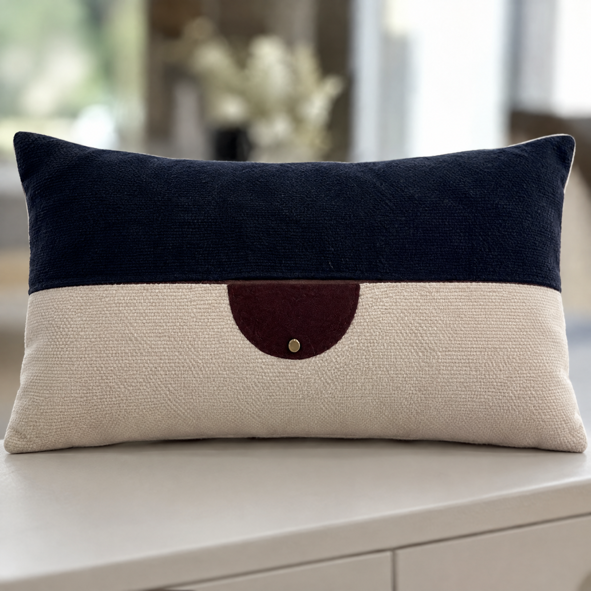 Close-up of a decorative pillow with a navy blue top section and a beige bottom section, featuring a dark red heart-shaped accent sewn onto the beige area, placed on a white surface with blurred background of flowers and natural light.