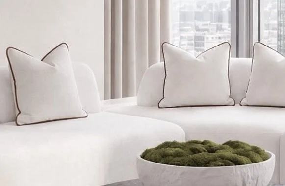 A white living room sofa with three white pillows, surrounded by sheer curtains and a city view outside the window, with a white bowl filled with green moss in front.