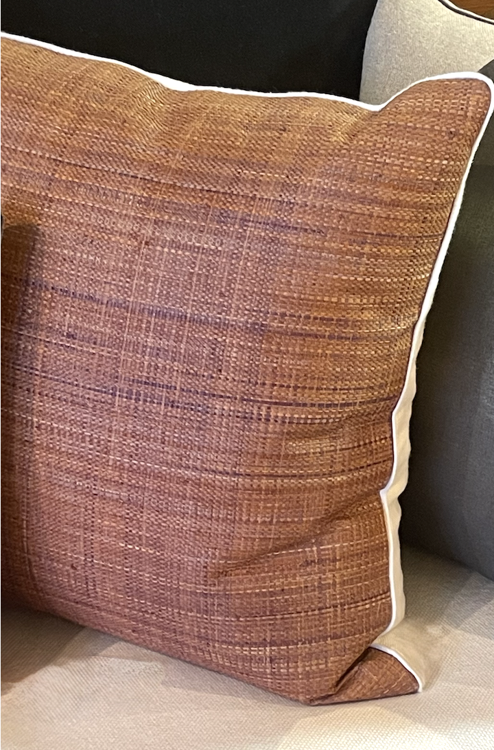 Close-up of a decorative throw pillow with a textured brown fabric cover and white piping, placed on a beige couch.