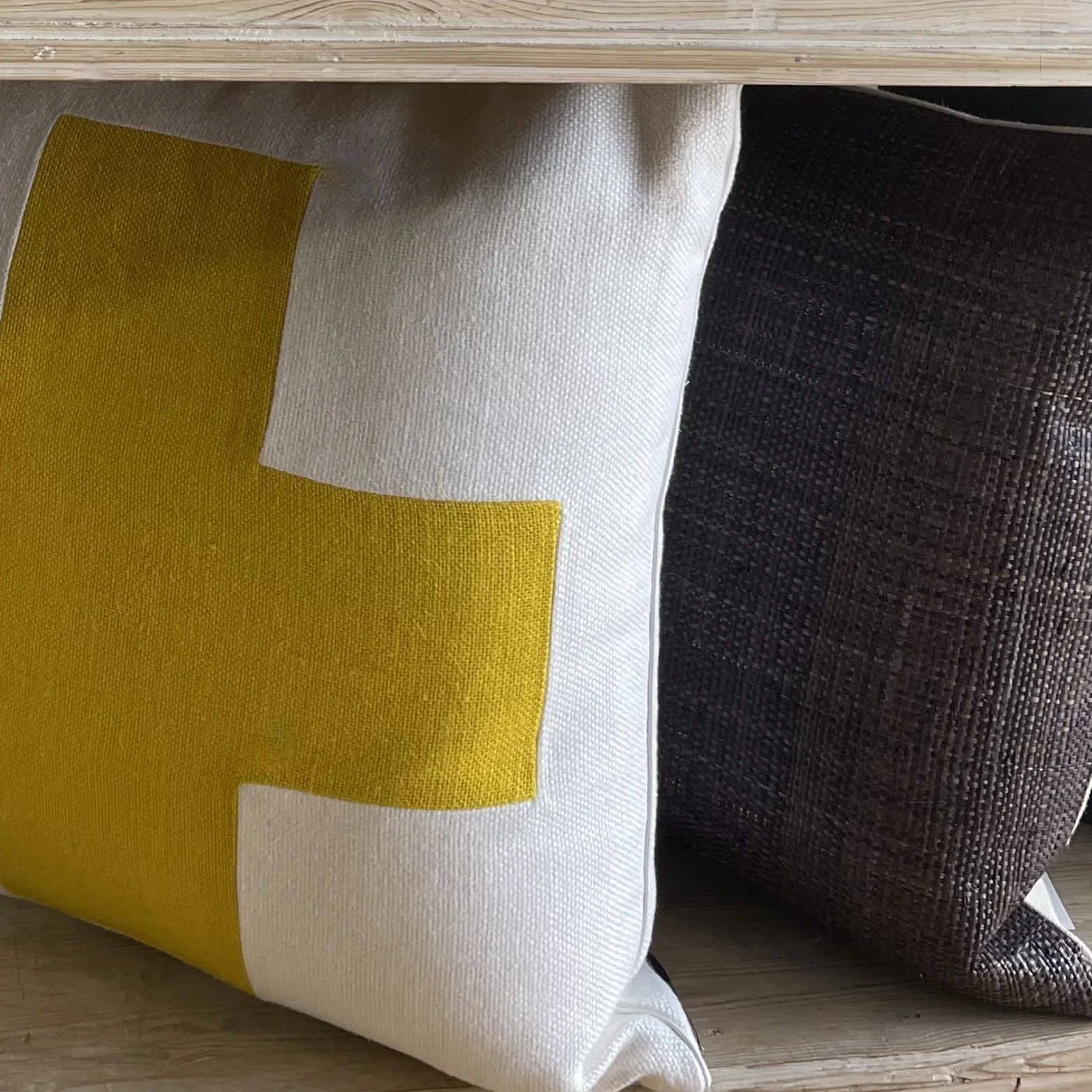Close-up of a fabric pillow with a yellow and white geometric pattern next to a dark brown textured pillow, placed on a light wooden surface.