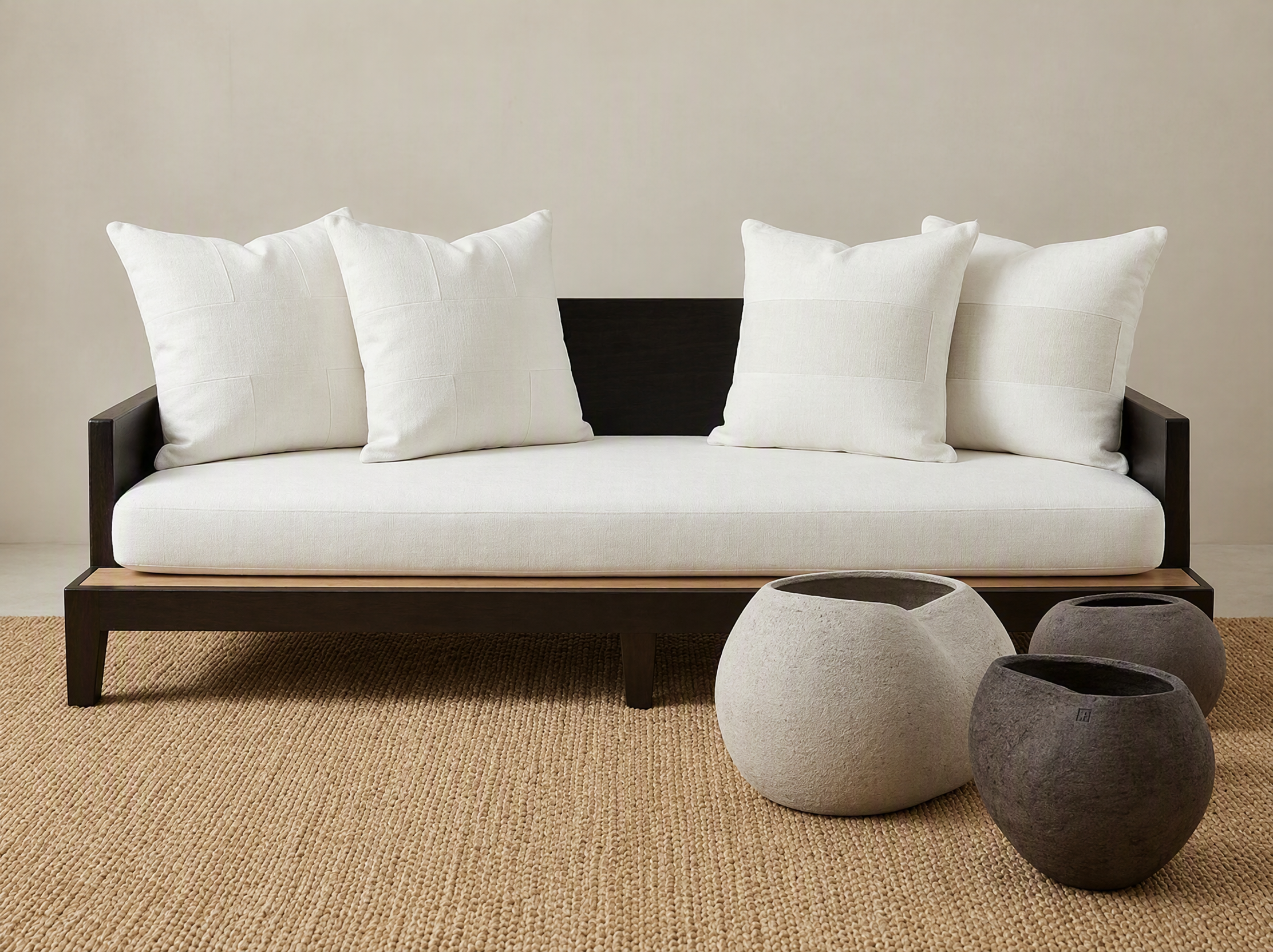 A contemporary living room setup with a white sofa adorned with three white pillows, set against a plain beige wall. In front of the sofa, there are three round, felted vases in gray, white, and black, placed on a woven beige rug.
