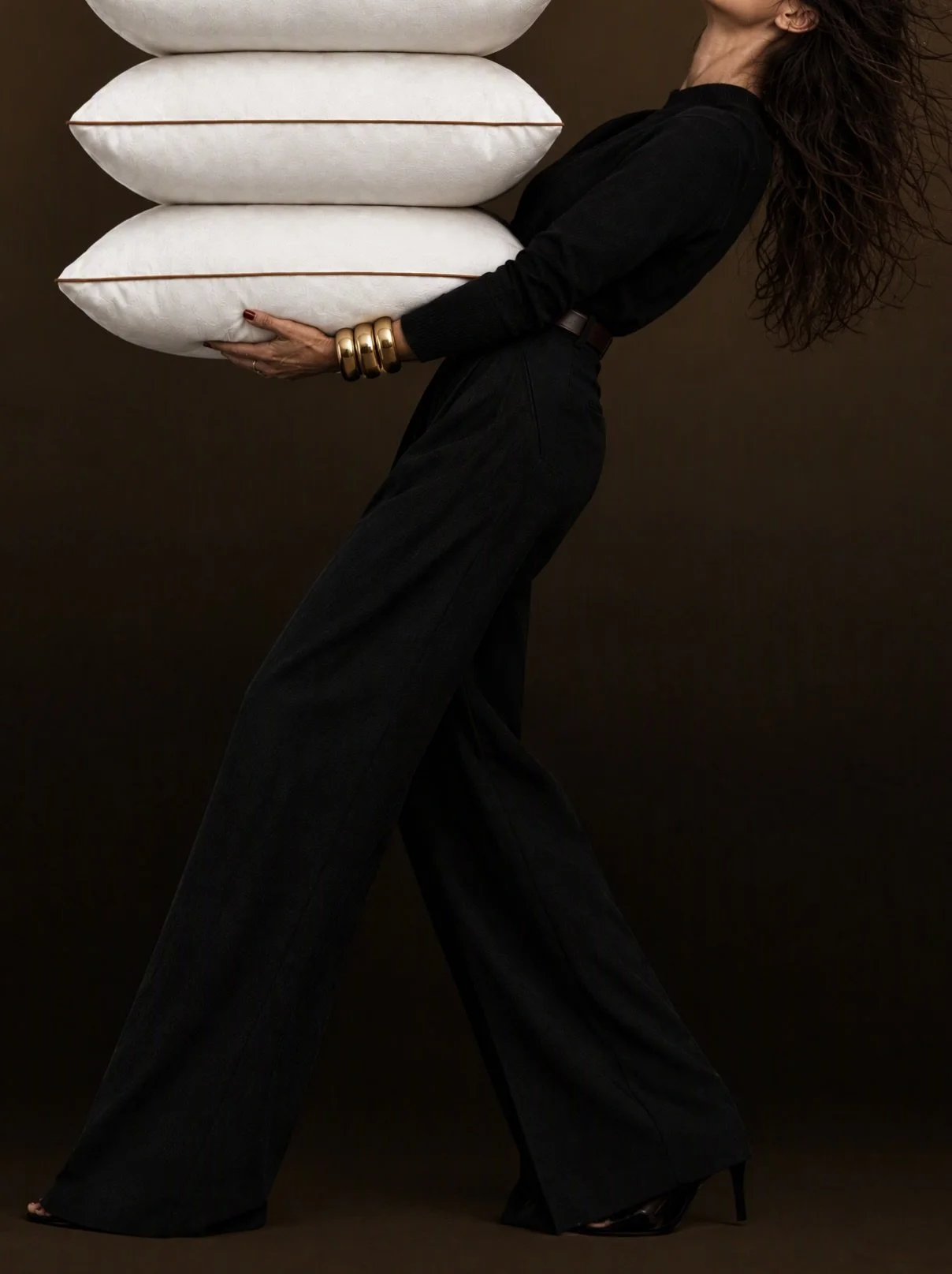 A woman with wavy hair wearing a black outfit and gold bangles, holding three stacked pillows against a dark background.