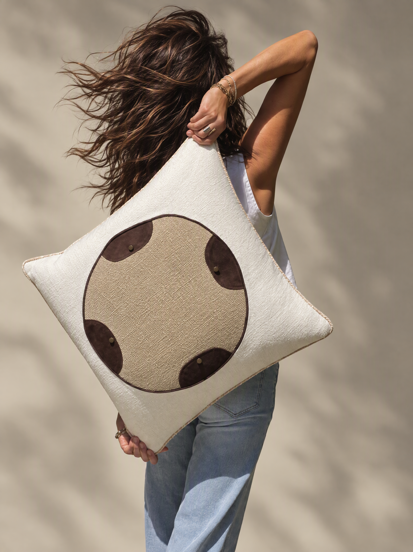 A person holding a large beige tote bag with a cartoon face design, standing against a blurred neutral background. The person has wavy brown hair, wears multiple rings and bracelets, and is dressed in a white top and light blue jeans.