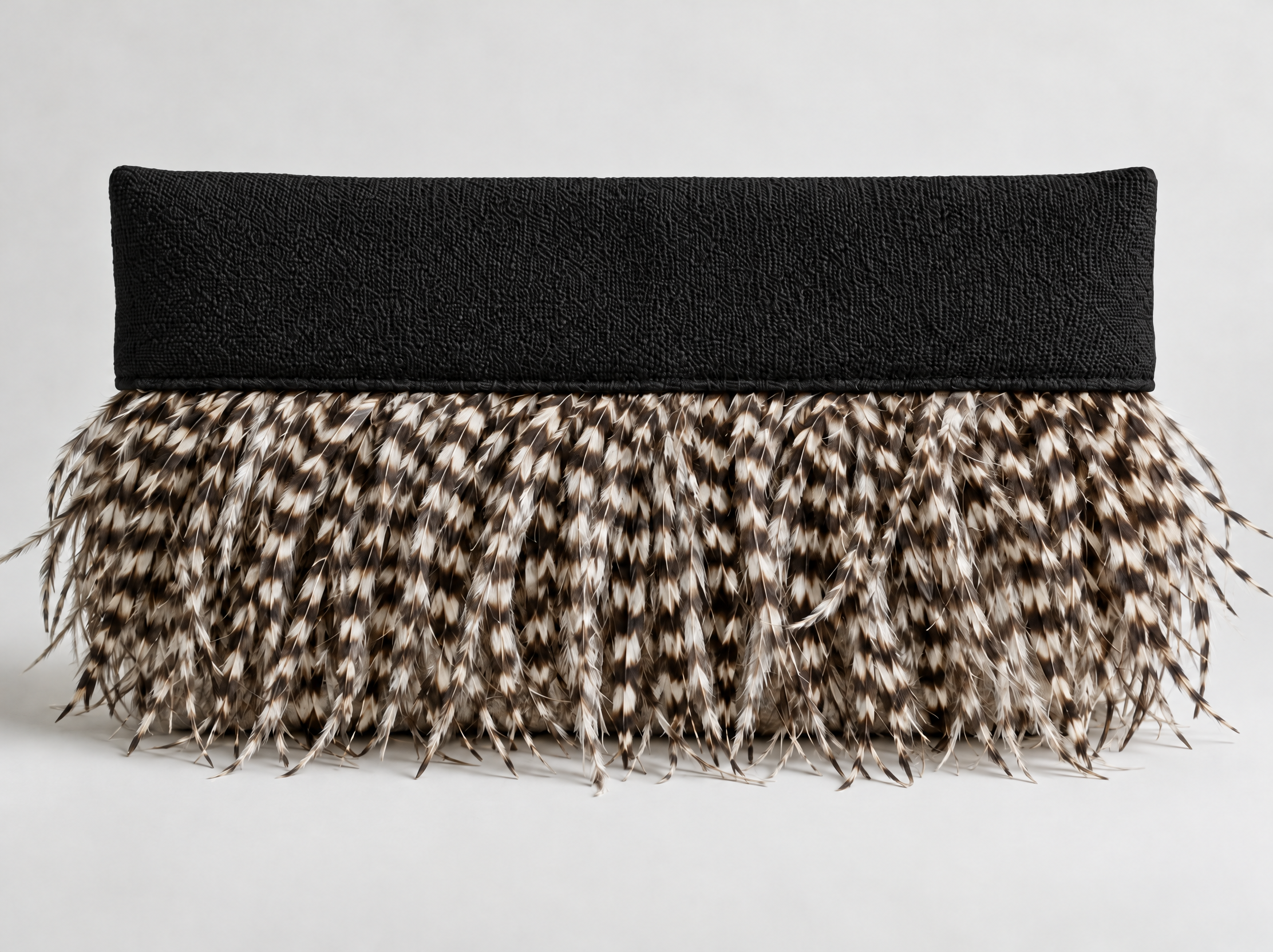 Close-up of a black cushion with textured fabric, sitting on a woven, spotted feathered surface, against a plain white background.