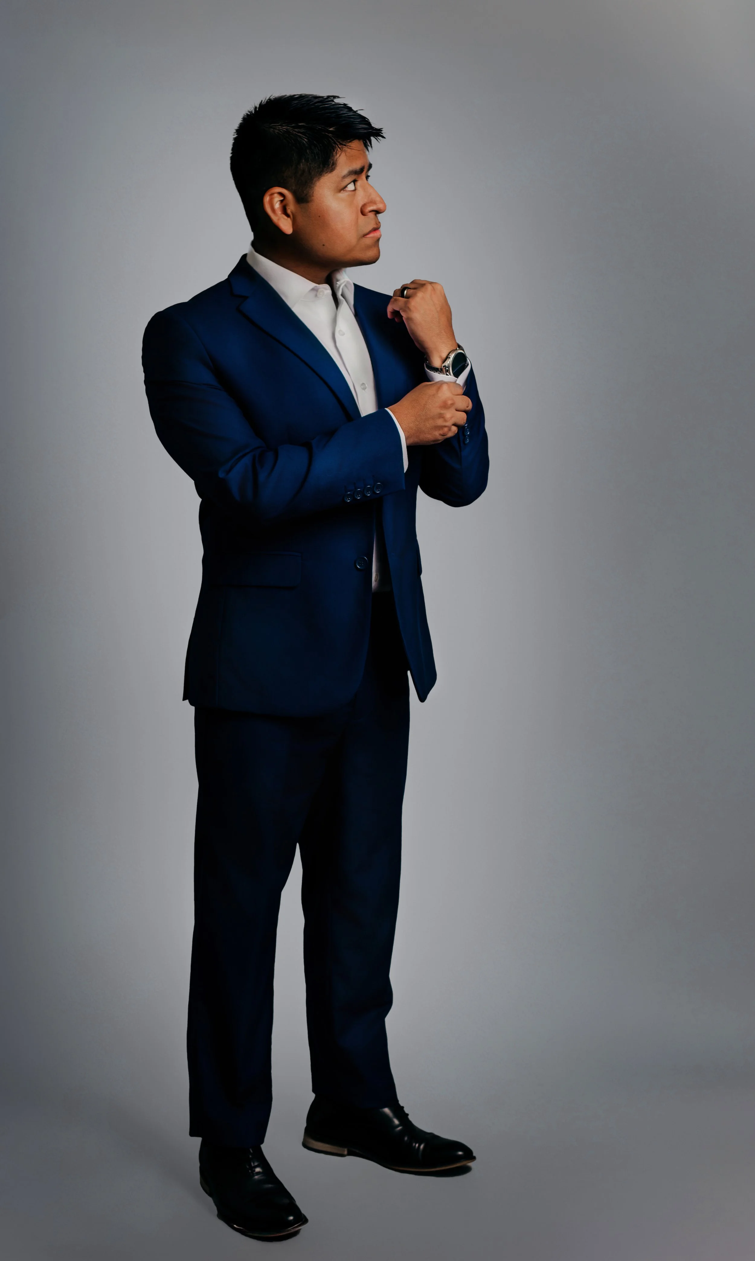 A man in a navy blue suit adjusting his watch against a plain gray background.