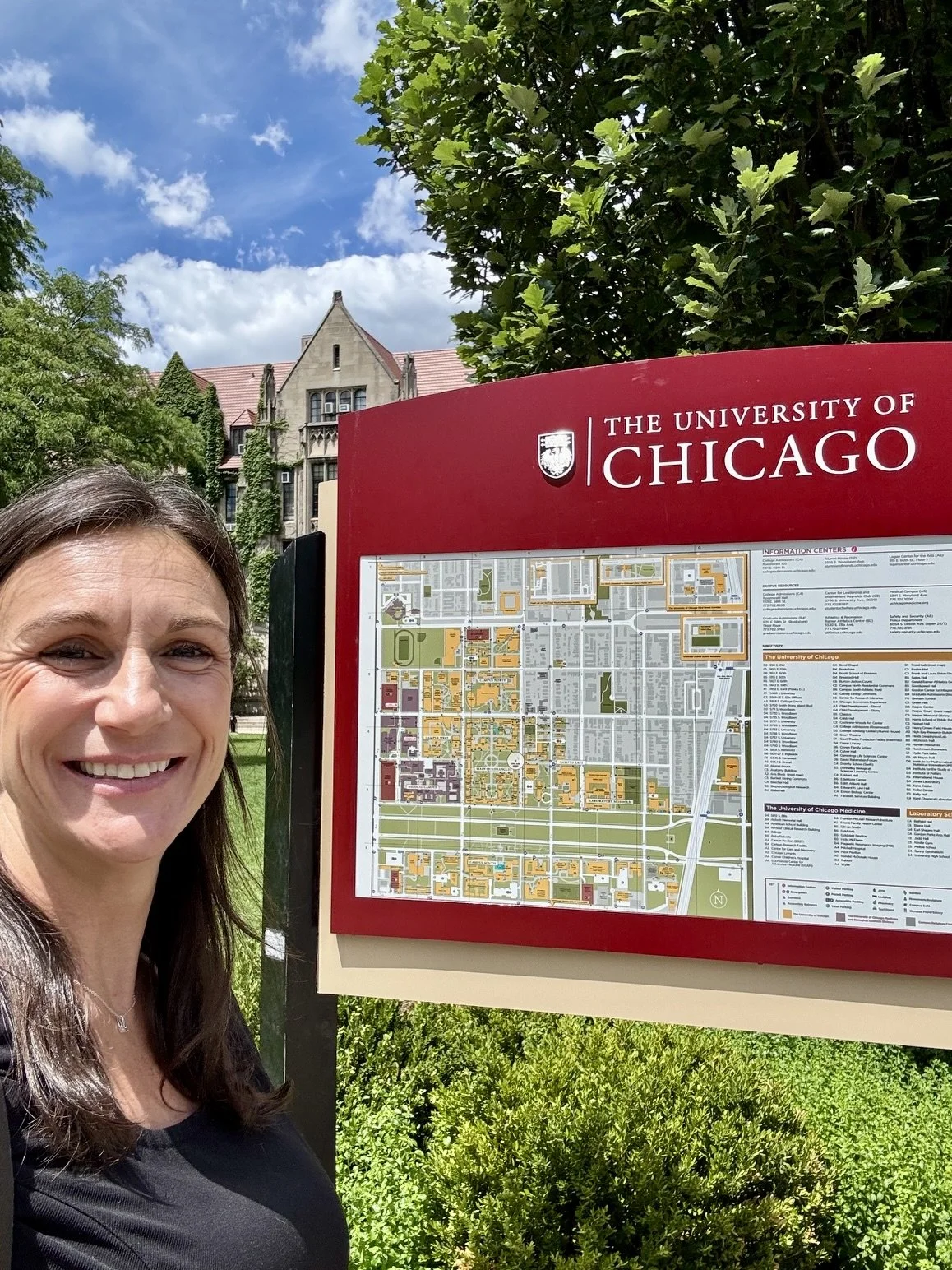 Smiling woman taking a selfie next to a campus map at The University of Chicago, with trees and historic buildings in the background on a sunny day.