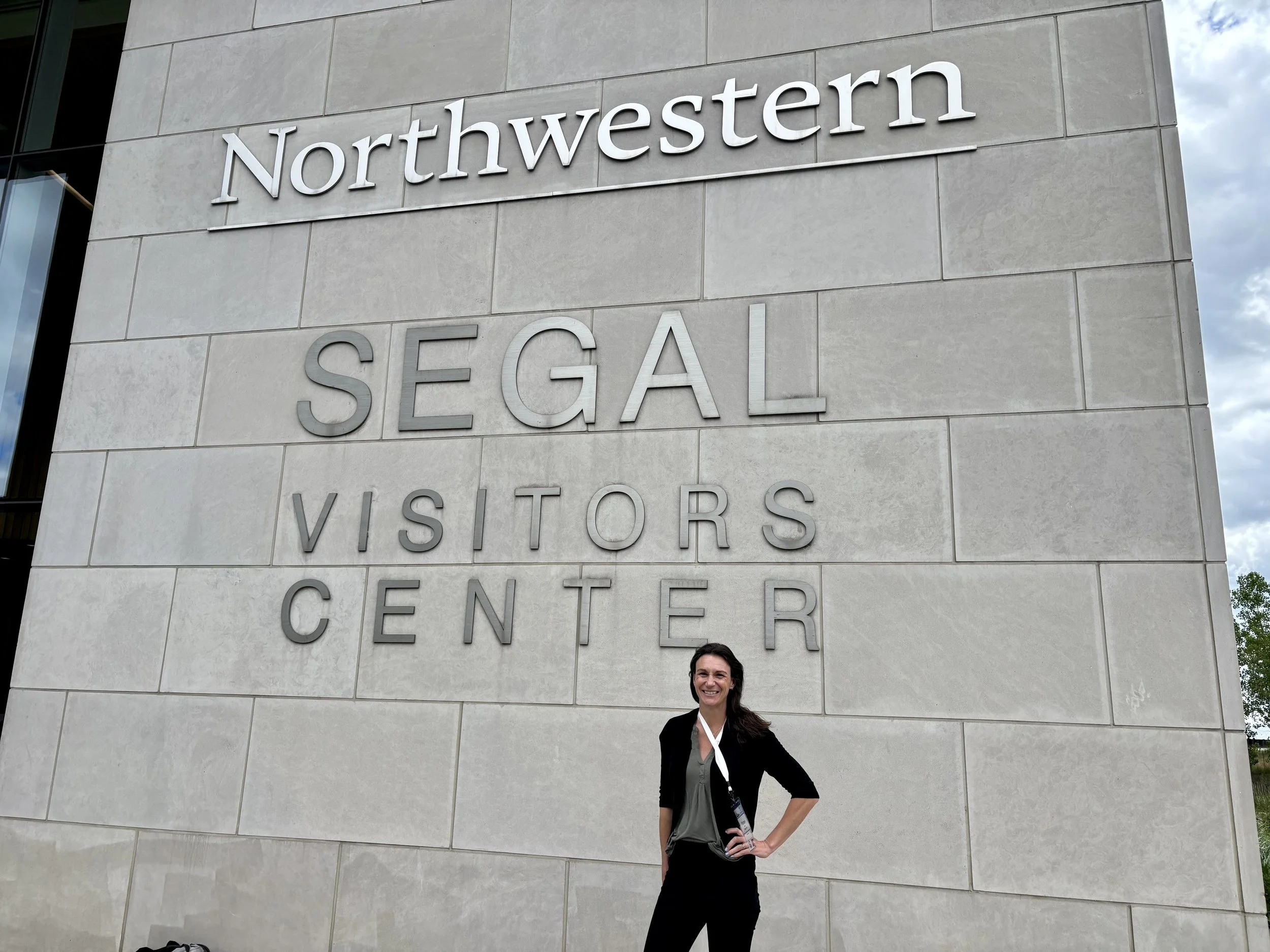 Photo of a smiling woman standing in front of a large building with the words 'Northwestern SEGAL' on the wall.