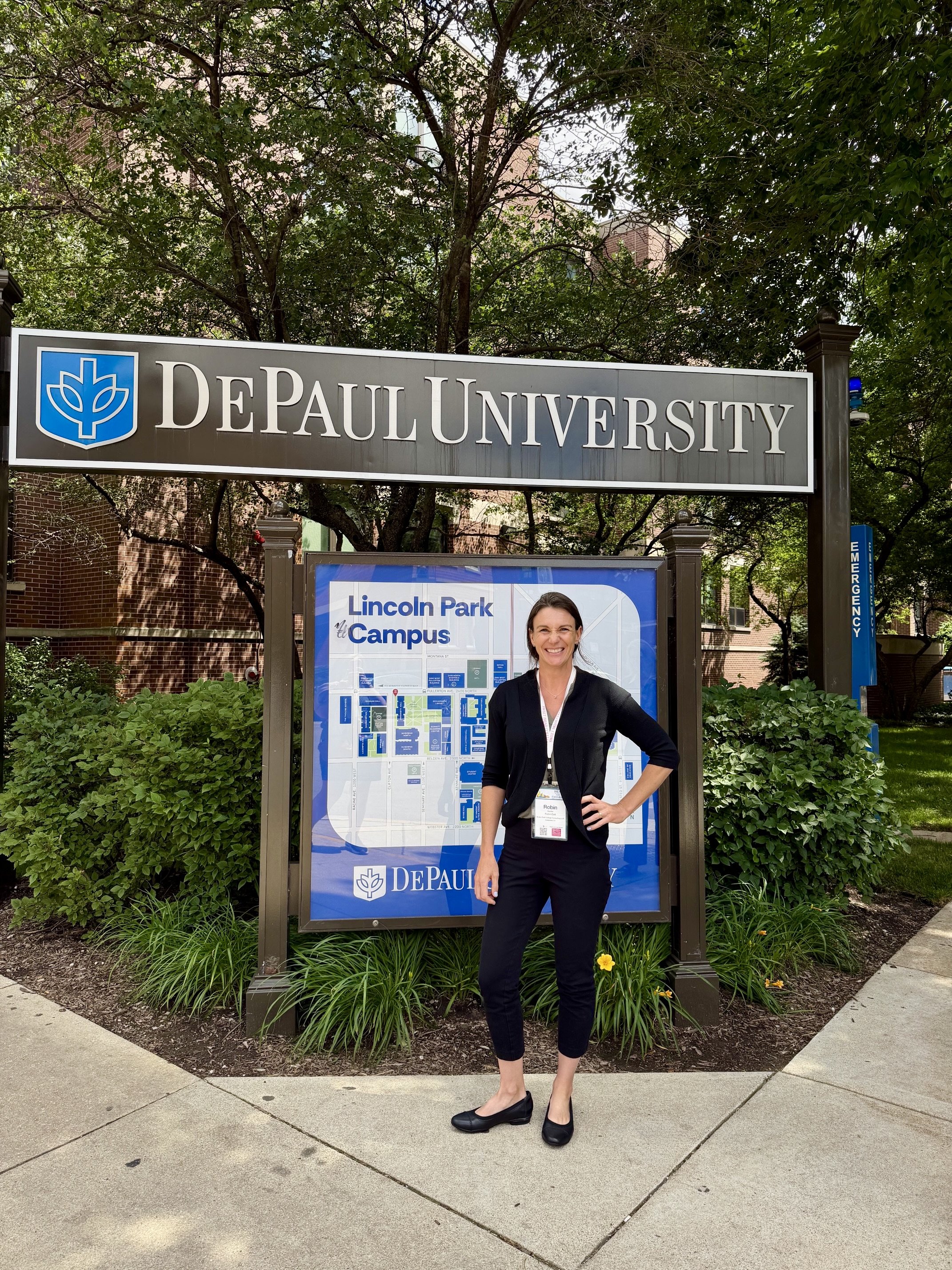 Woman standing in front of DePaul University sign on Lincoln Park Campus, smiling, wearing black blazer, black pants, and black shoes, with greenery and informational signage behind her.