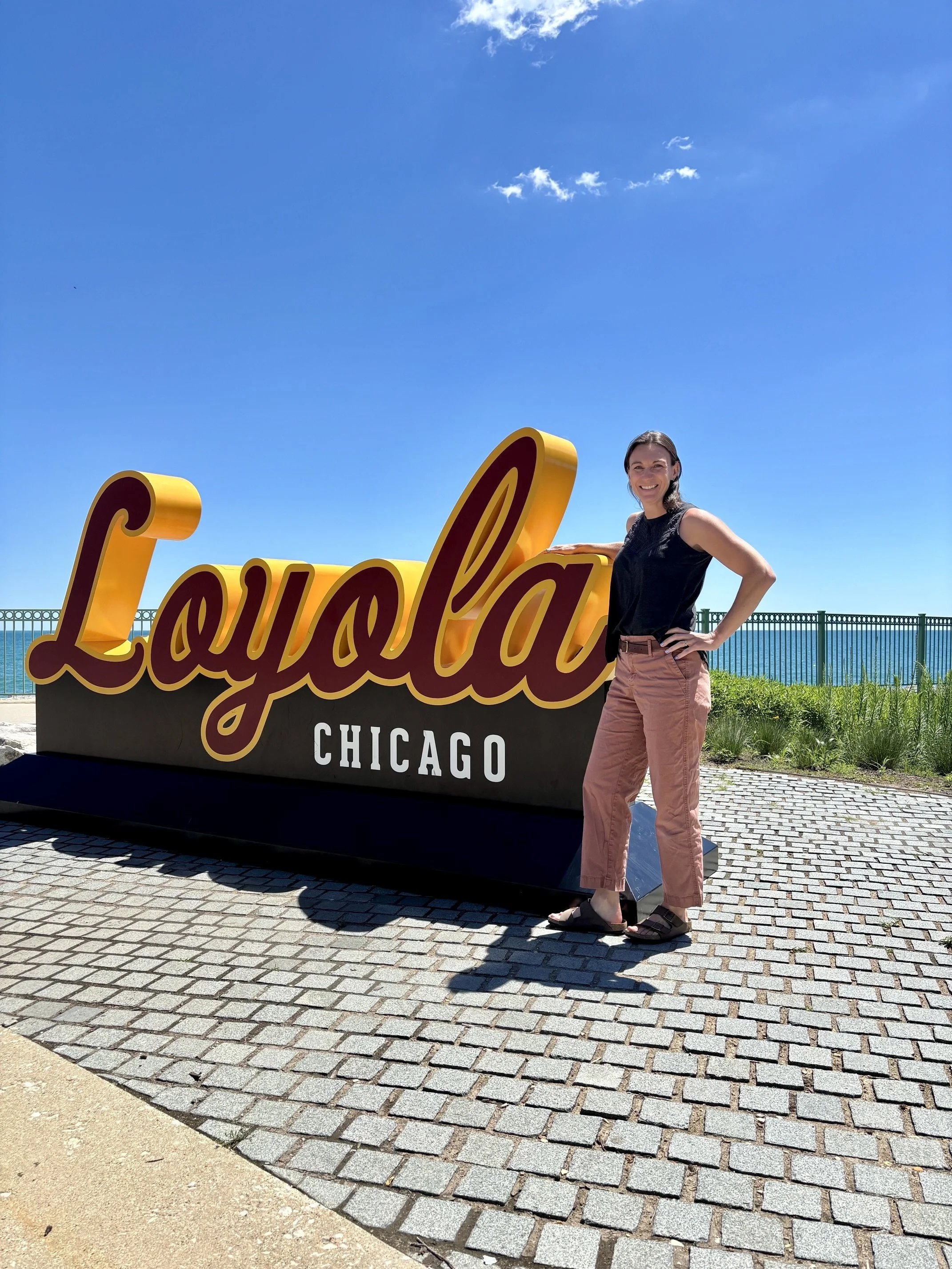 A woman standing next to a large sign that says "Chicago" in cursive letters, with a blue sky and a few clouds in the background. She is smiling, wearing a black top, pink pants, and sandals, with one hand on her hip and the other resting on the sign