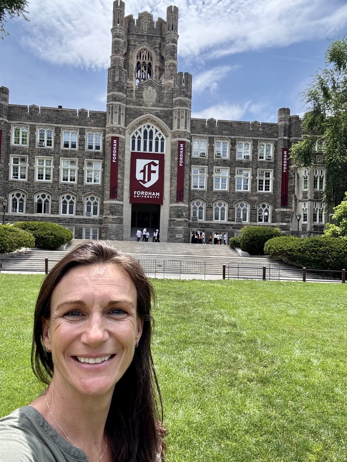 A woman smiling in front of Fordham University’s Gothic-style main building, with stairs, greenery, and students in the background on a sunny day.