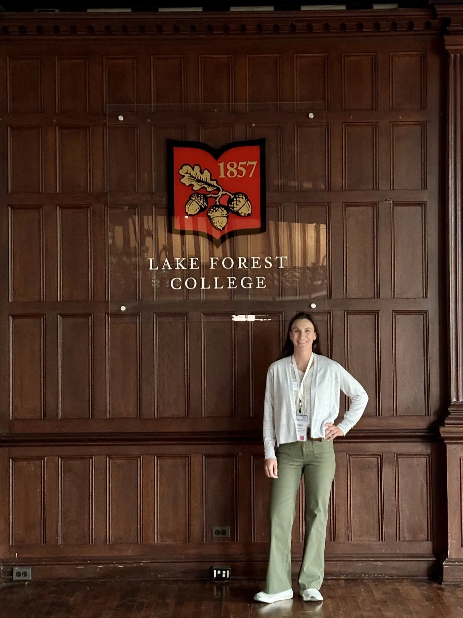 A woman standing in front of a wooden wall with a sign that reads 'Lake Forest College' and a logo with acorns and the year 1857. She is wearing a white jacket, green pants, and white shoes, and has a visitor badge around her neck.