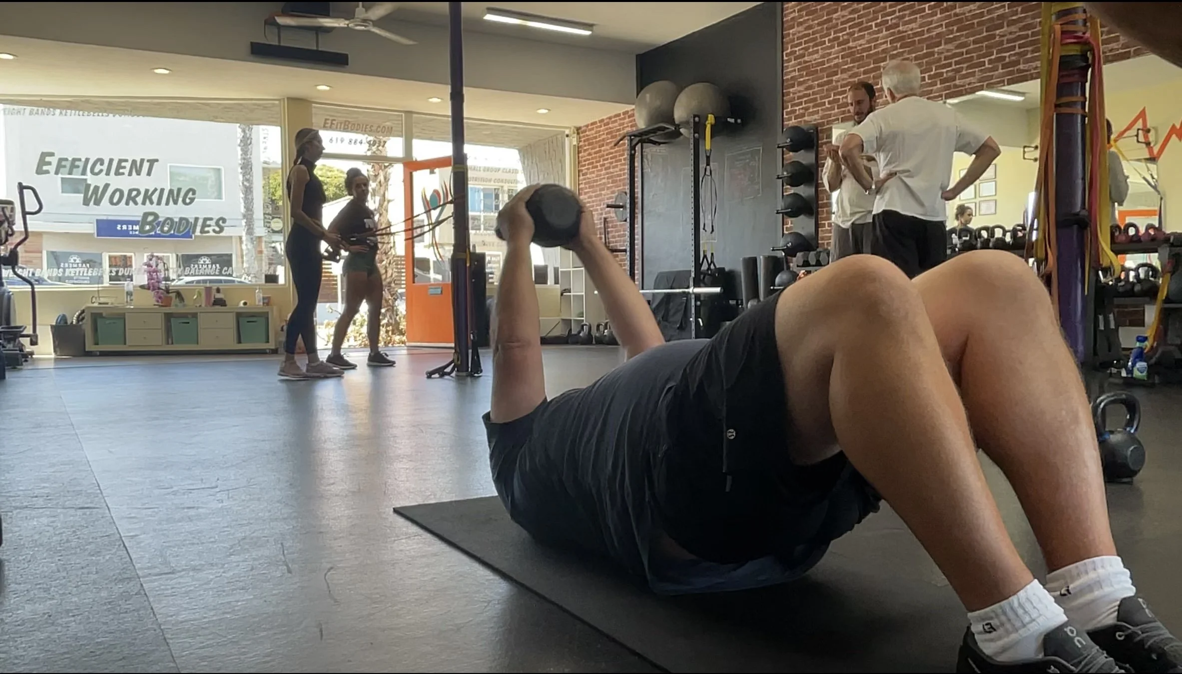 A person lying on a workout mat lifting a dumbbell in a gym with people working out and talking in the background.