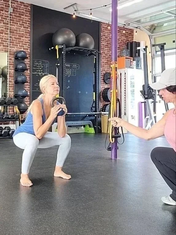 A woman in workout clothes performing a squat with a kettlebell in a gym, assisted by a trainer holding resistance bands in front of her, with exercise equipment and a brick wall in the background.