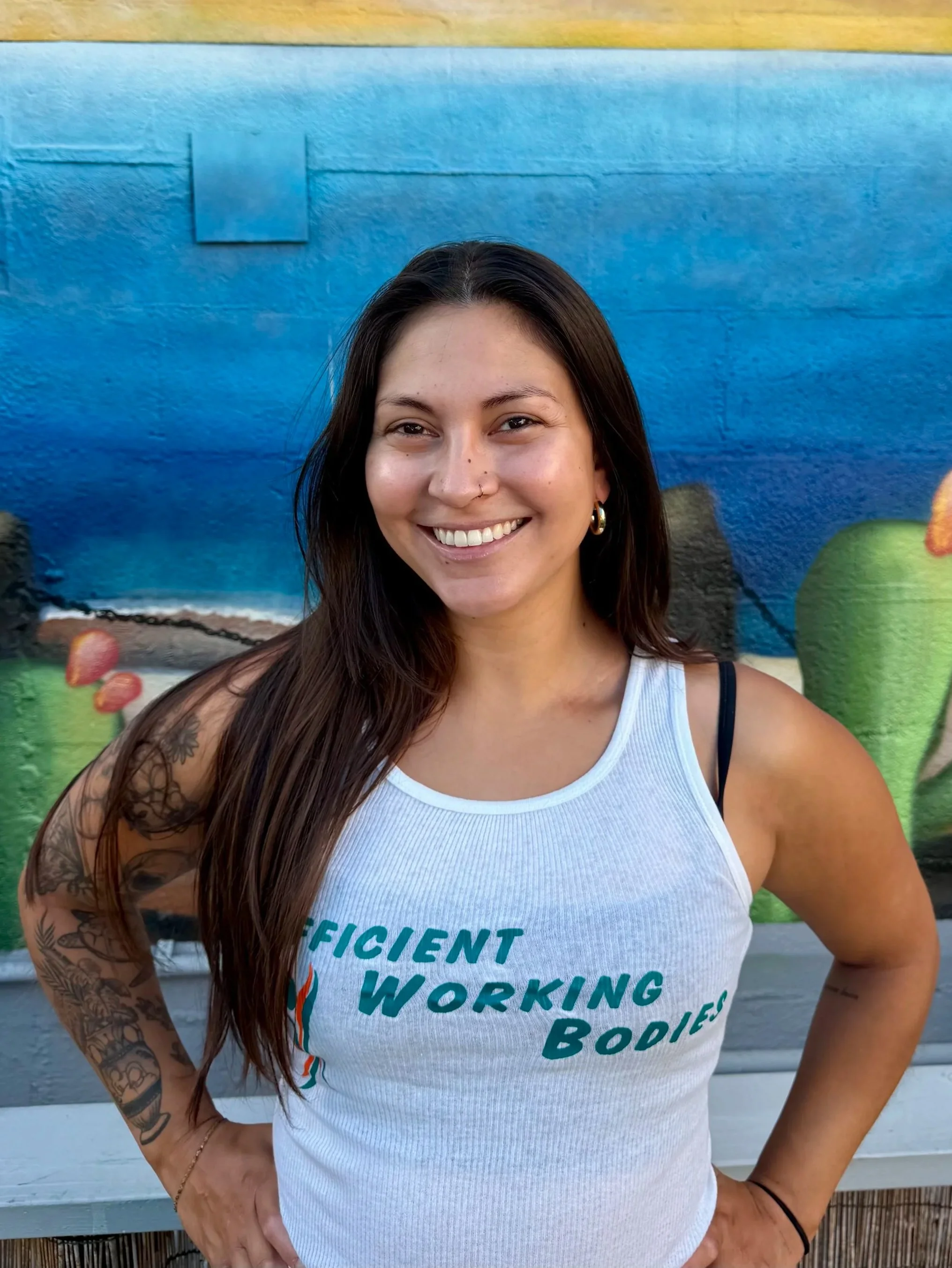 A woman with long, dark hair and tattoos on her left arm, smiling with her hands on her hips in front of a colorful mural with blue water, green cacti, and strawberries.