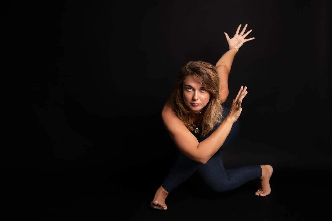 Woman performing a yoga pose against a black background, wearing dark clothing and looking into the camera.