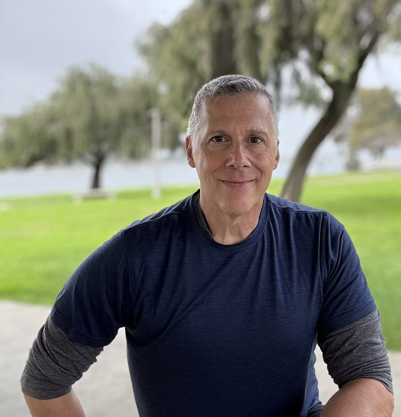 A middle-aged man with short gray hair, smiling, wearing a navy blue t-shirt with gray sleeves, outdoors in a park with green grass, trees, and a body of water in the background.