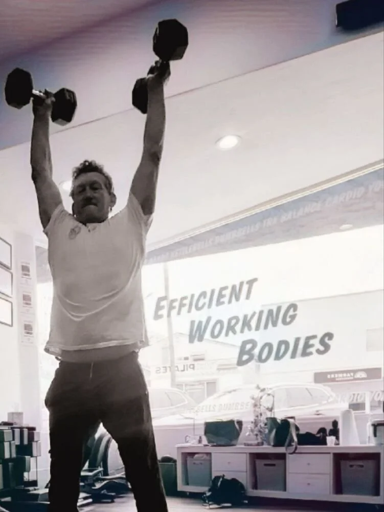 A man lifting dumbbells overhead in a gym with a window behind him, displaying the words 'Efficient Working Bodies' and a street scene outside.