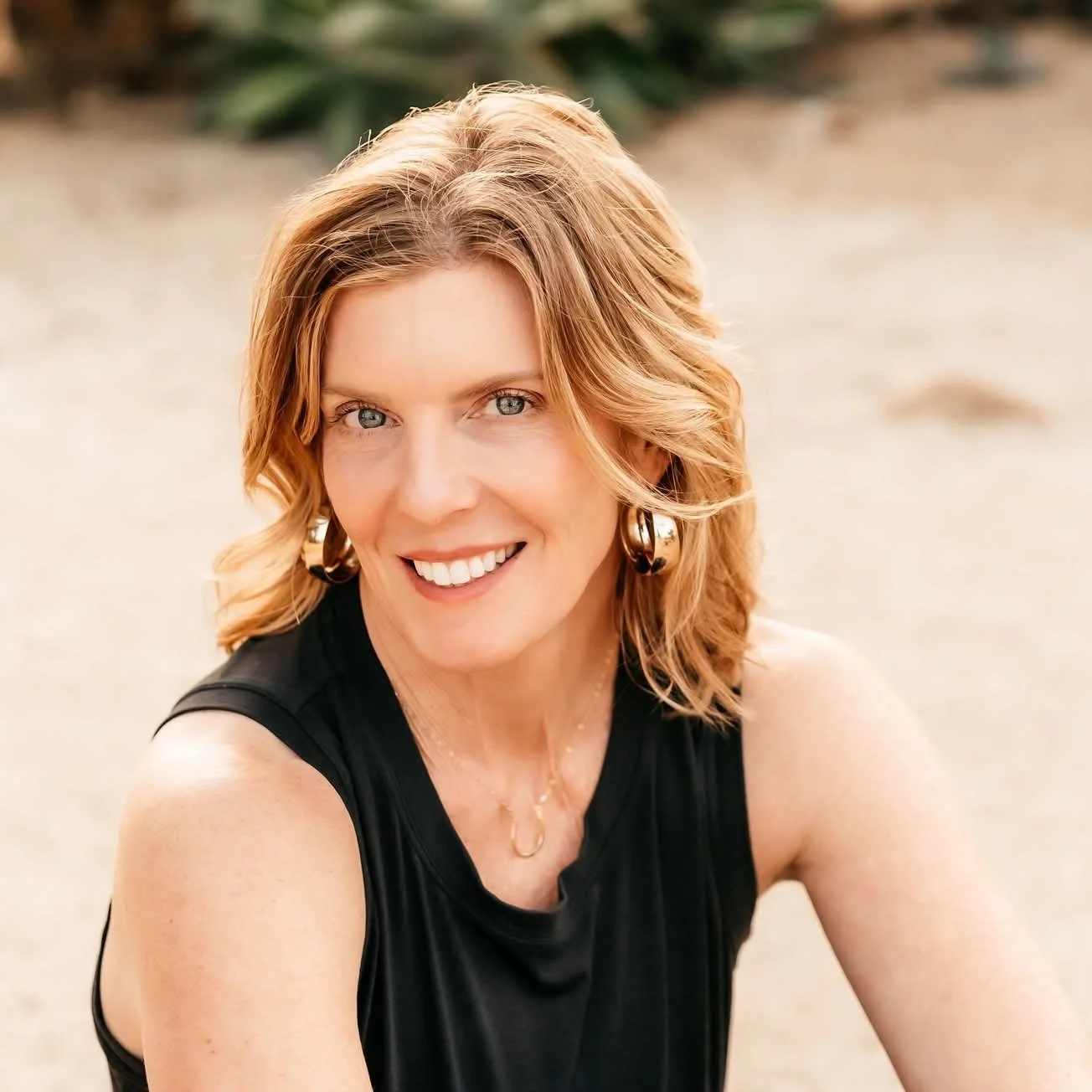 A woman with shoulder-length wavy red hair and blue eyes, smiling and wearing a sleeveless black top with gold jewelry, outdoors on a sandy beach.