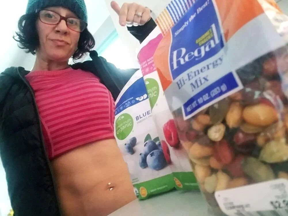 Woman with glasses, wearing a striped pink top and a beanie, lifts her shirt to reveal her stomach and pose with a smile. In the foreground, there are bags of organic trail mix and a cereal box on a table.