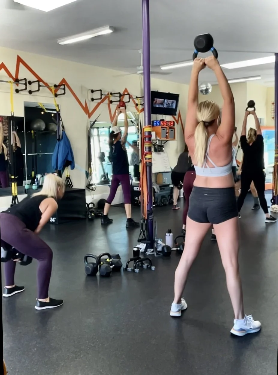 Women exercising with kettlebells and dumbbells in a gym, with fitness equipment and a red zigzag line design on the wall.