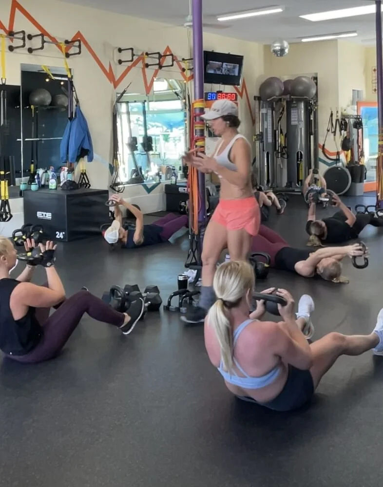 People exercising in a gym using kettlebells, lying on the floor, and sitting on the floor. A fitness instructor is standing in the center, instructing the class.