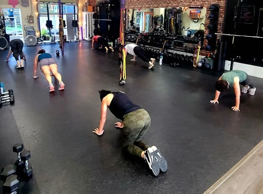 People participating in a group workout class in a gym, doing push-ups and other exercises on a black rubber floor.