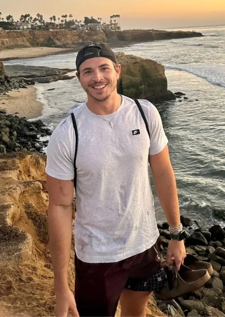 A smiling young man wearing a black cap, white t-shirt, and shorts standing on a rocky shoreline at sunset with the ocean, cliffs, and palm trees in the background.