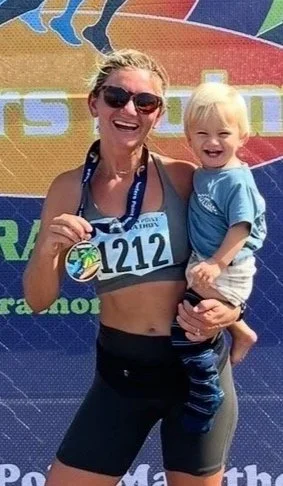A woman in athletic gear holding a young blonde girl, both smiling, with a colorful event backdrop behind them.