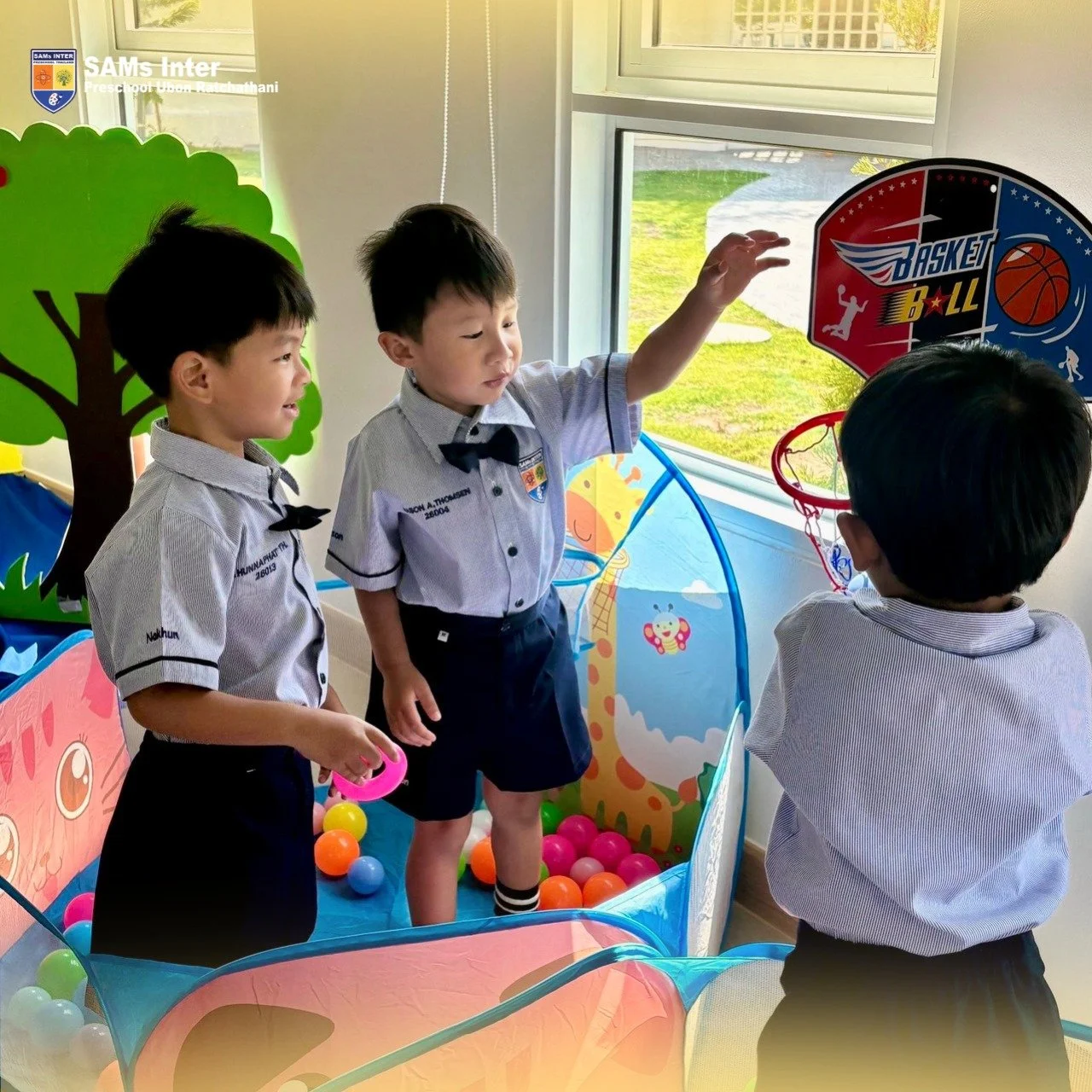 Three young boys in school uniforms playing basketball in an indoor play area with a ball pit, colorful walls, and a window showing a grassy outdoor area.