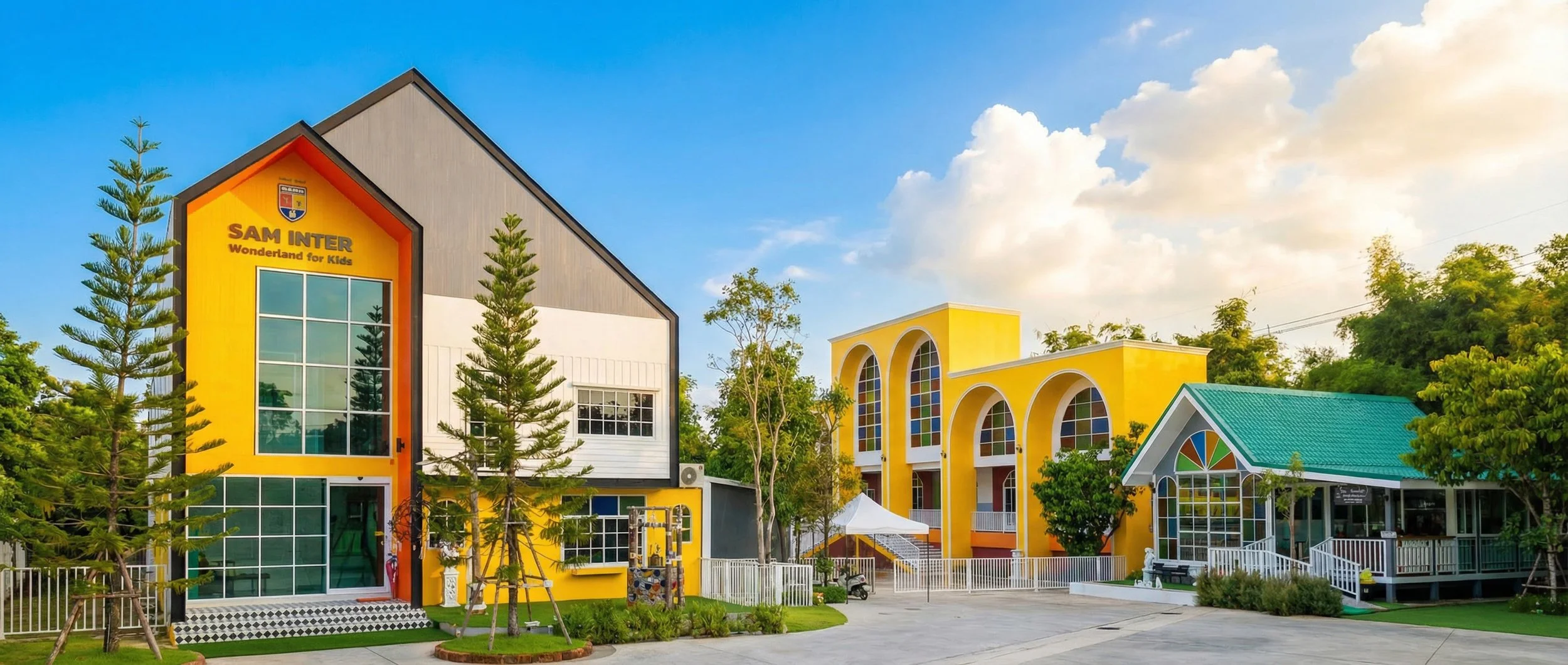 Colorful educational building with large windows and yellow walls, surrounded by green trees, under a partly cloudy sky.