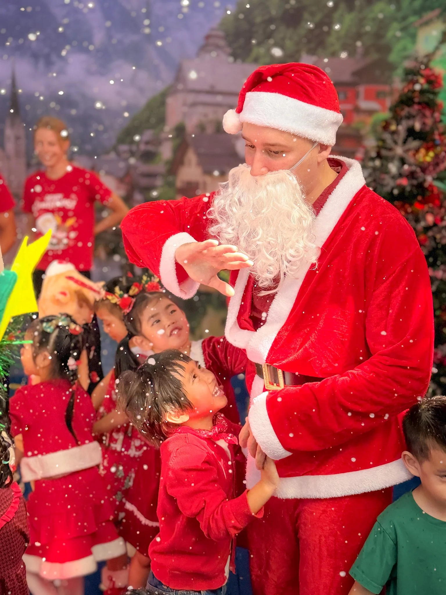 A man dressed as Santa Claus is surrounded by children, some of whom are reaching up to him. The man is wearing a red Santa suit with white fur trim, a Santa hat, and a fake white beard. It is snowing. In the background, there is a Christmas tree and a painted backdrop of a townscape.
